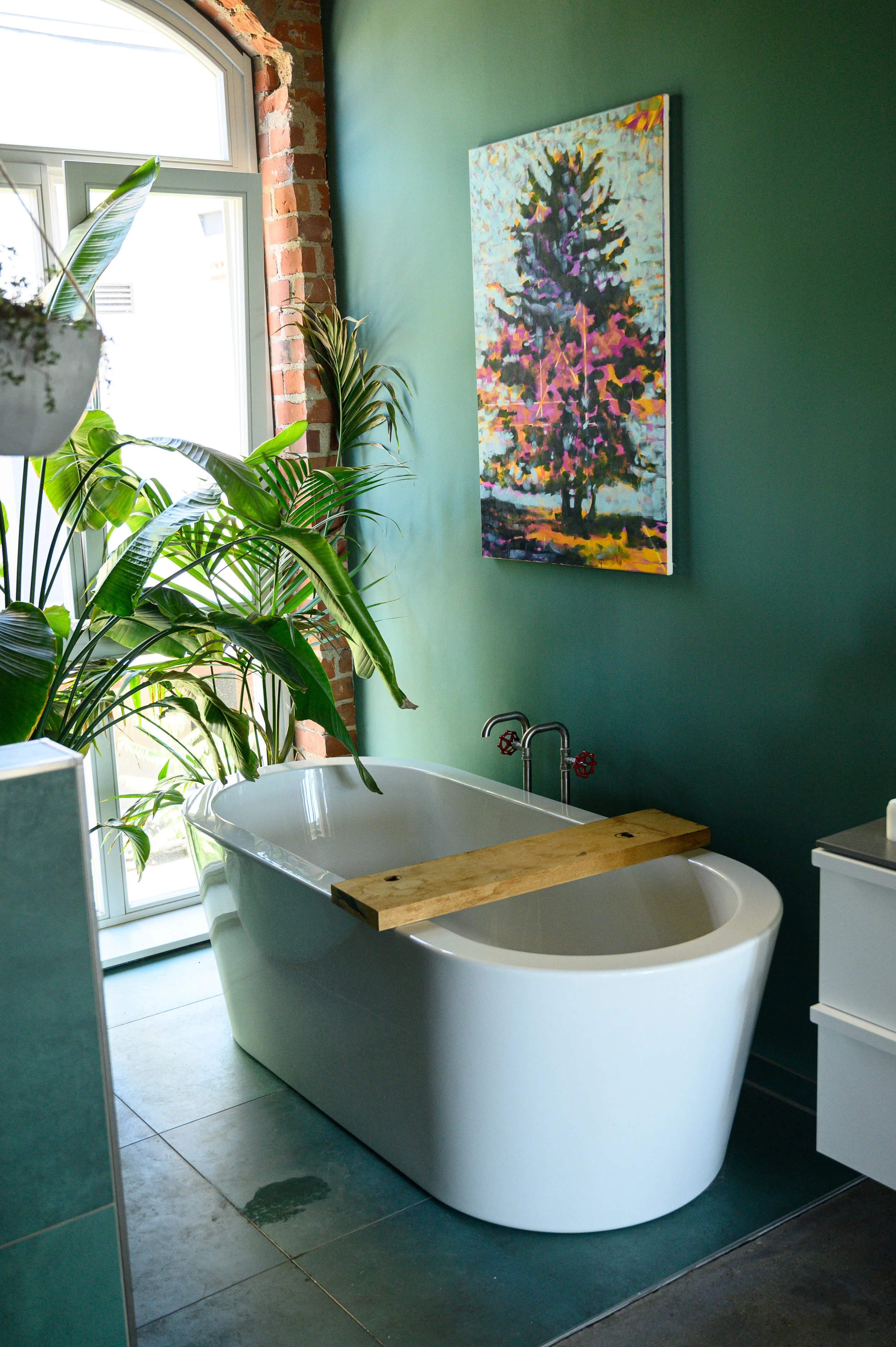 Ein modernisiertes Badezimmer mit freistehender Badewanne, grüner Wandfarbe, Pflanzen an einem Fenster, und einem bunten Baum-Gemälde an der Wand.