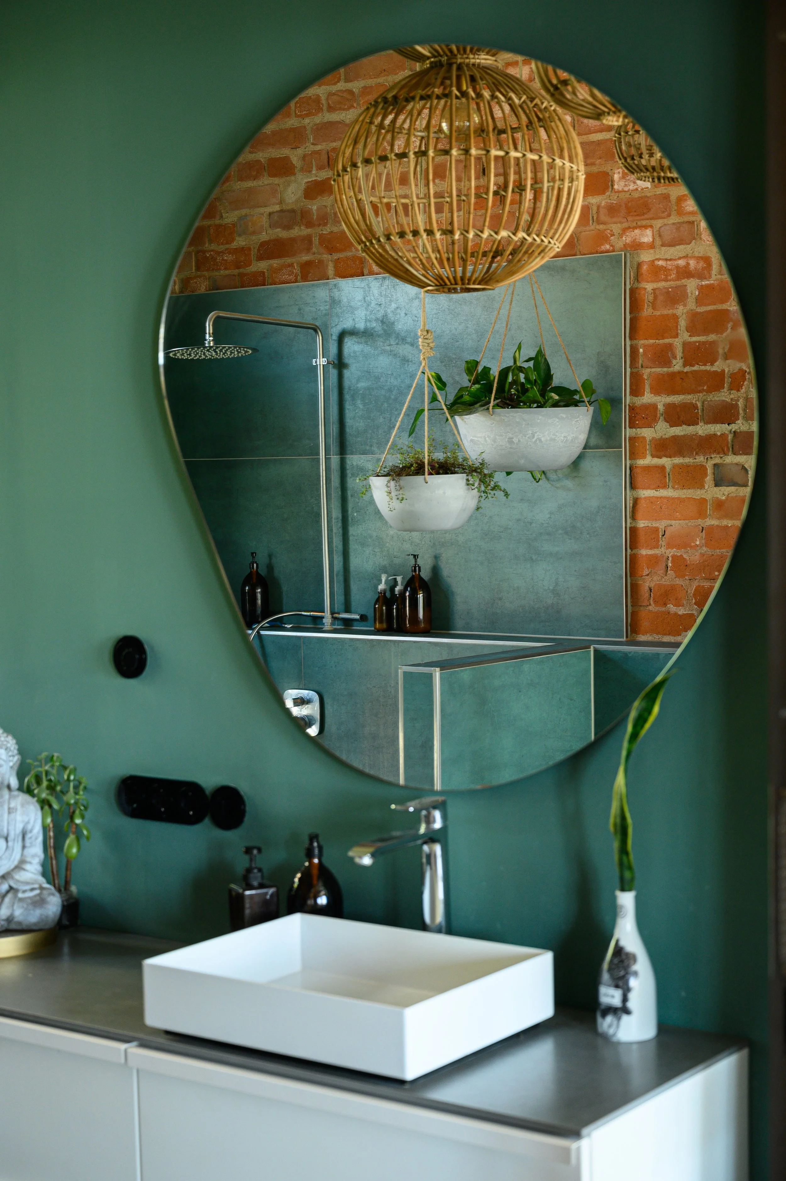 Ein Badezimmer mit grünem Wand, Spiegel, Waschbecken und Pflanzen, gesehen durch einen unregelmäßig geformten Spiegel.