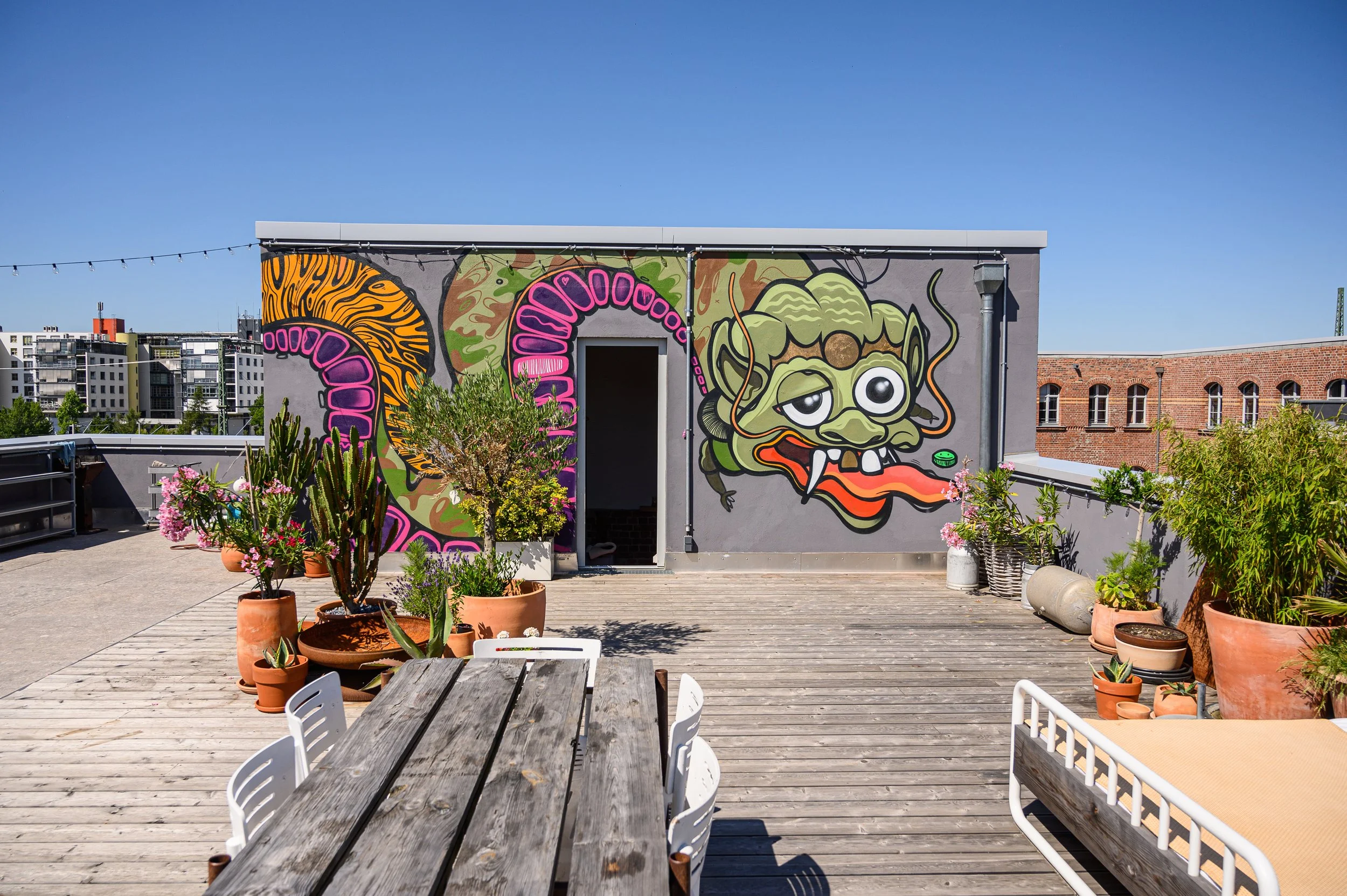 Dachterrasse mit buntem Graffiti eines Drachen auf grauer Wand, Gartenpflanzen in Tontöpfen, Holztisch und Stühlen bei sonnigem Himmel.