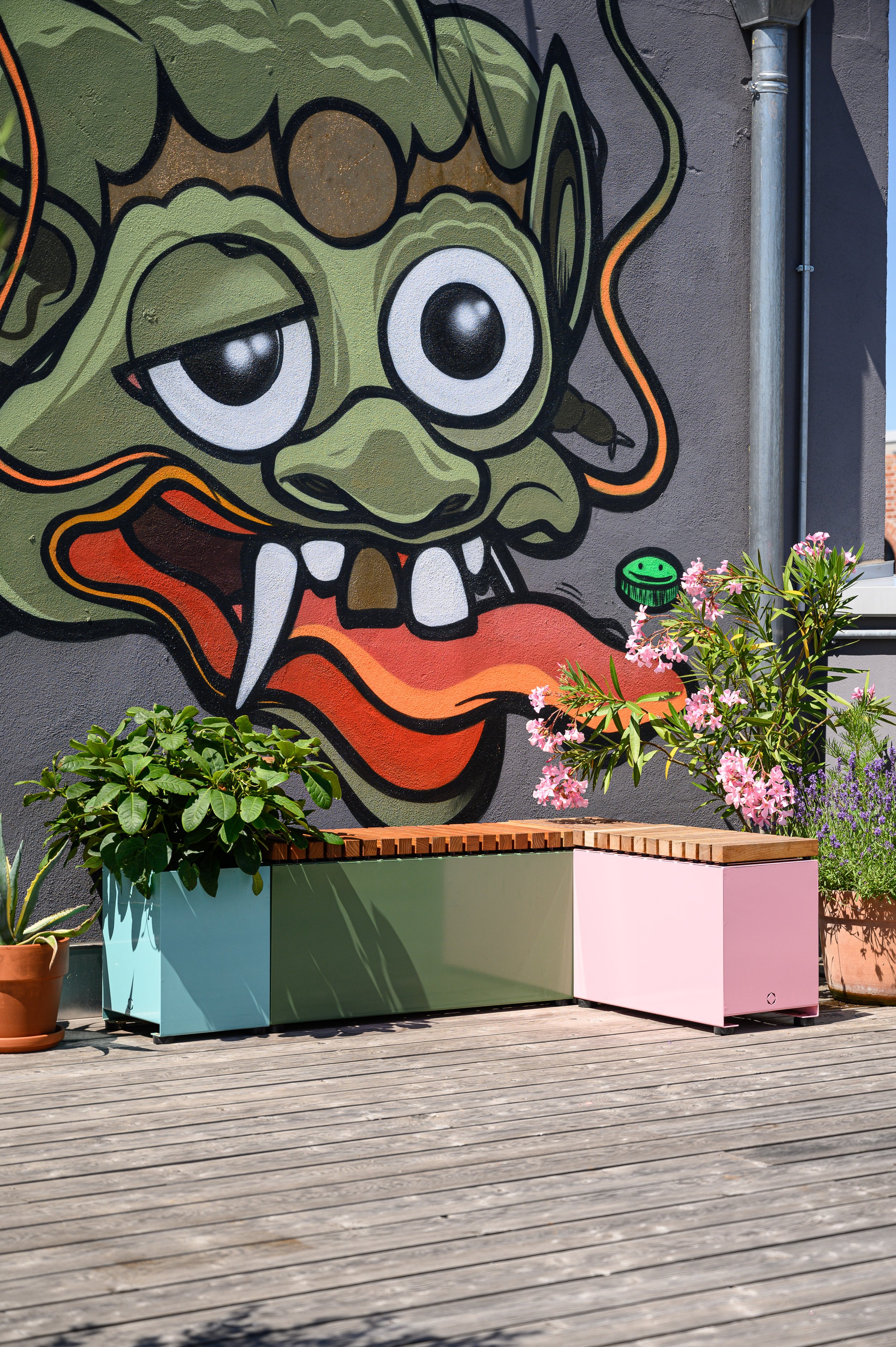 Street Art eines grünen, dämonischen Kreatur mit großen Augen und Zähnen, umgeben von Pflanzen auf einem Balkon mit Holzboden.