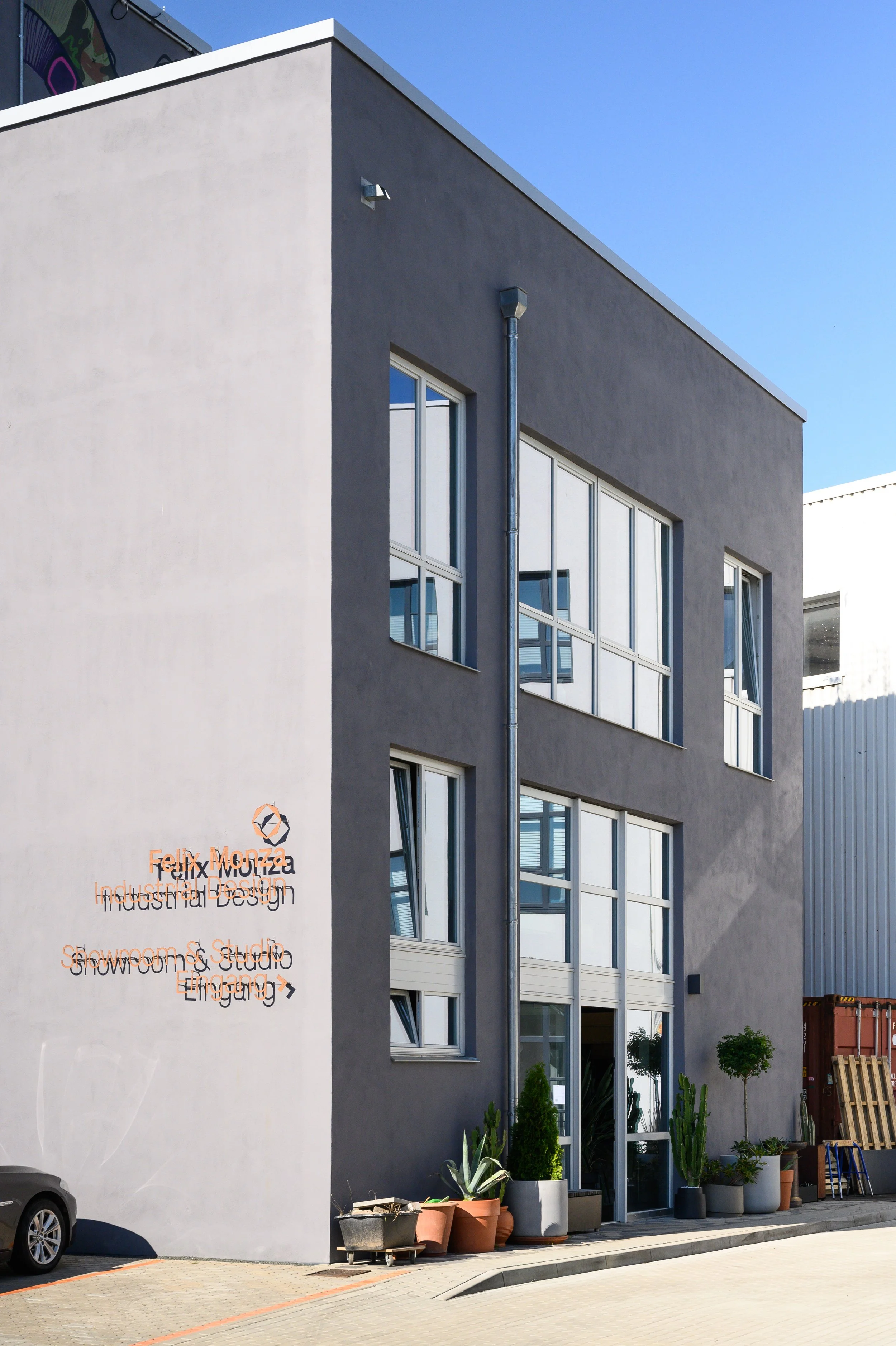 Das moderne, mehrstöckige Gebäude mit grau verputzter Fassade und großen Fenstern. Am Eingang befinden sich diverse Topfpflanzen und eine Beschilderung mit dem Text 'Felix Monza, Industrial Design, Snowoom & Studio, Einstieg'.