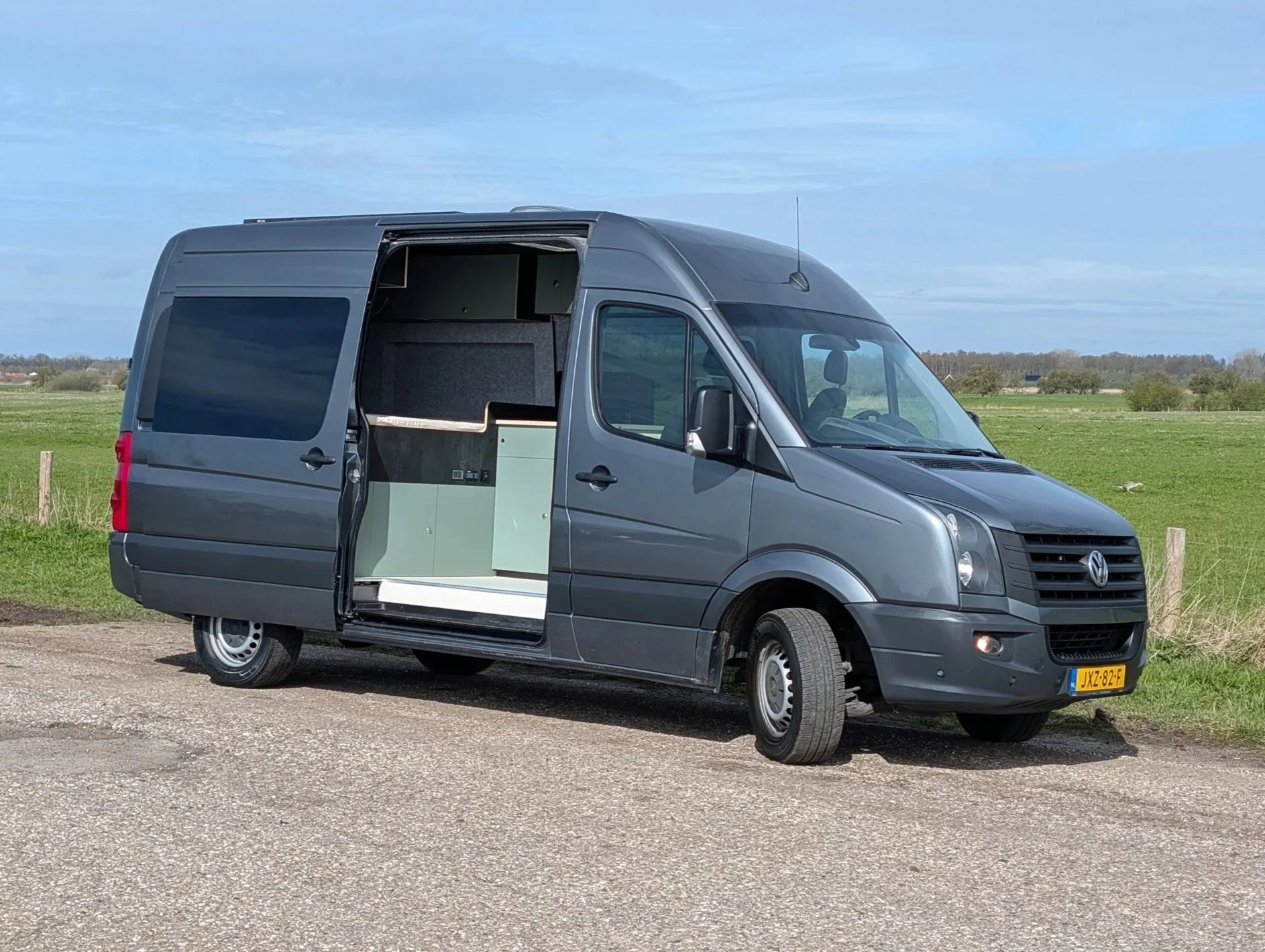 Een grijze Volkswagen Camperbus op een landweg, met open schuifdeur die de binnenkant toont.