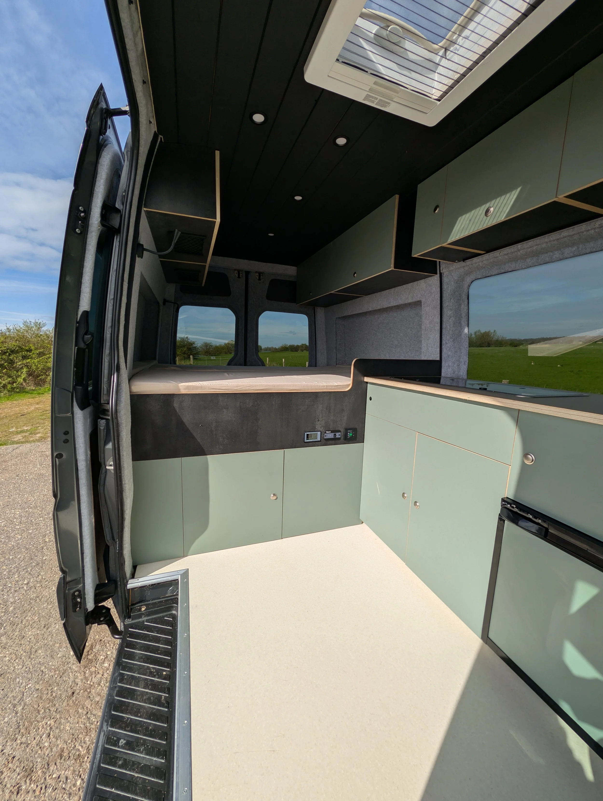 Interieur van een camper met een kitchenette en een bed, uitzicht op groene velden en een blauwe lucht buiten.