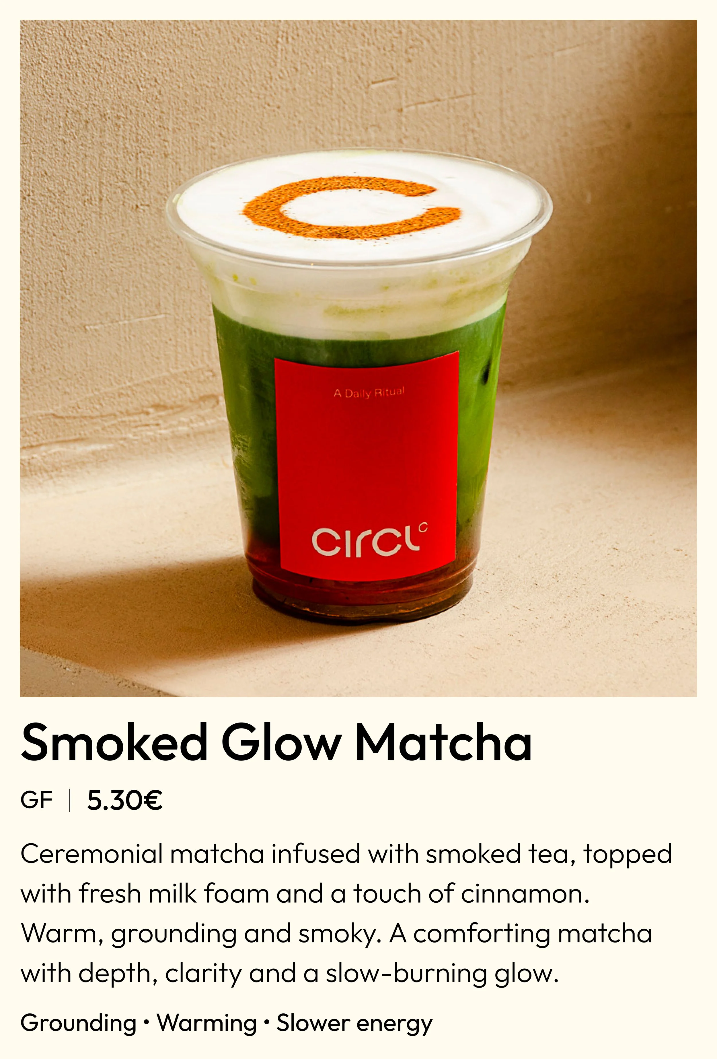 A cup of Smoked Glow Matcha with a cinnamon 'C' on top of the foam, placed on a neutral surface against a beige wall.