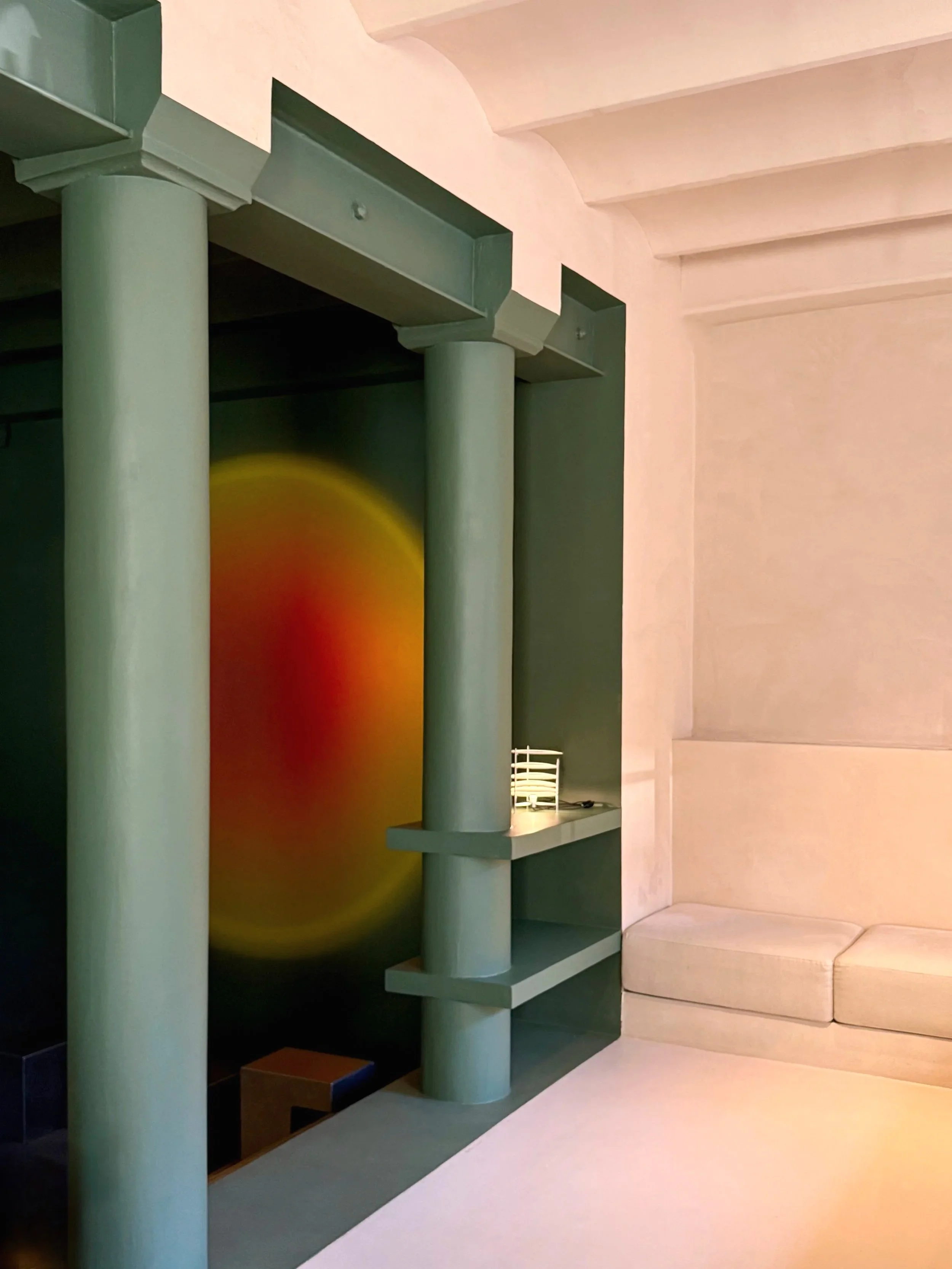 Interior space with cream-colored wall, green columns, and built-in shelving, featuring a colorful abstract painting with red and yellow hues.