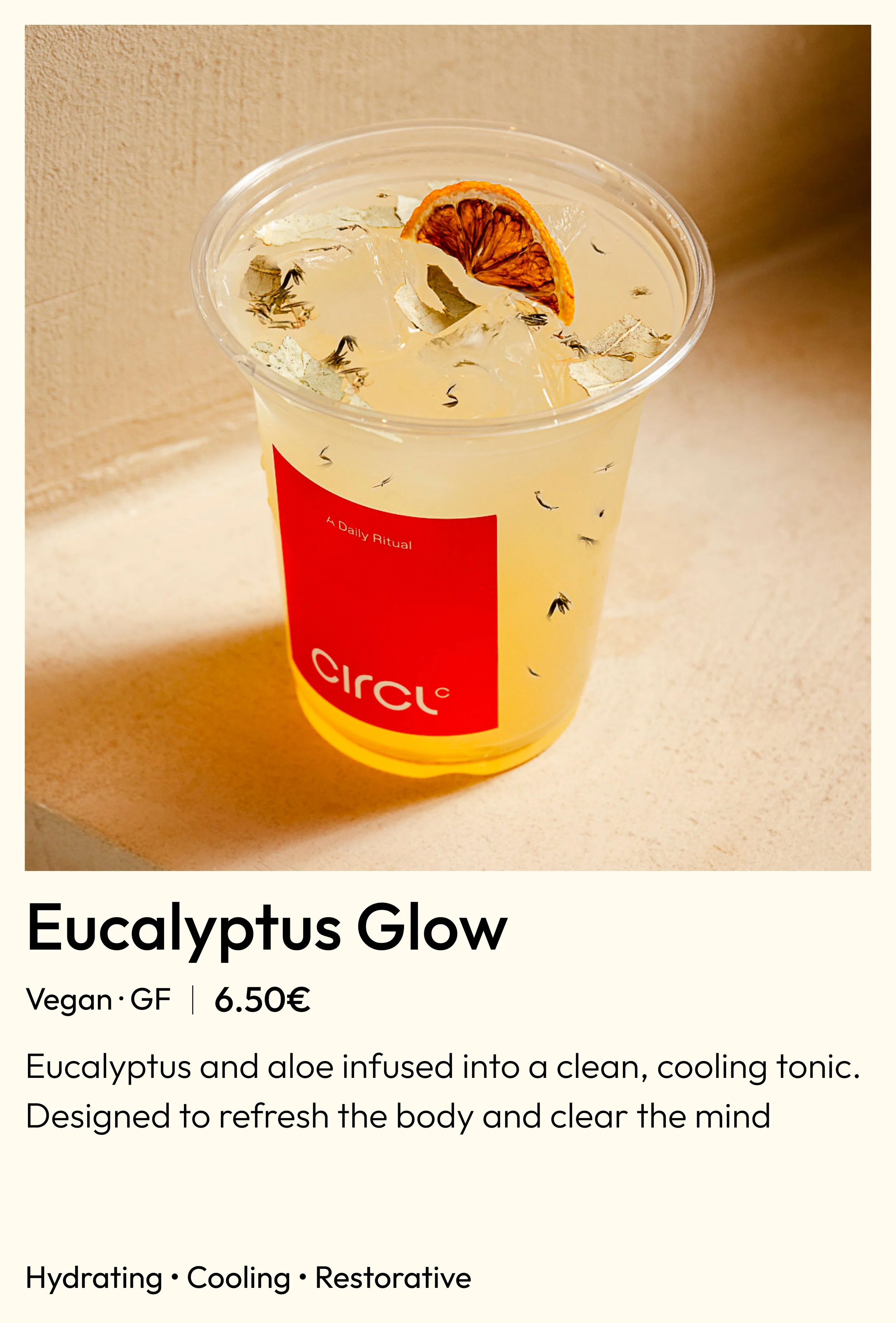 A clear plastic cup filled with a light yellow beverage, ice cubes, dried orange slices, and herbs. The cup has a red label with the word 'circ' and the text 'A Daily Ritual' printed on it. The background is a beige surface.
