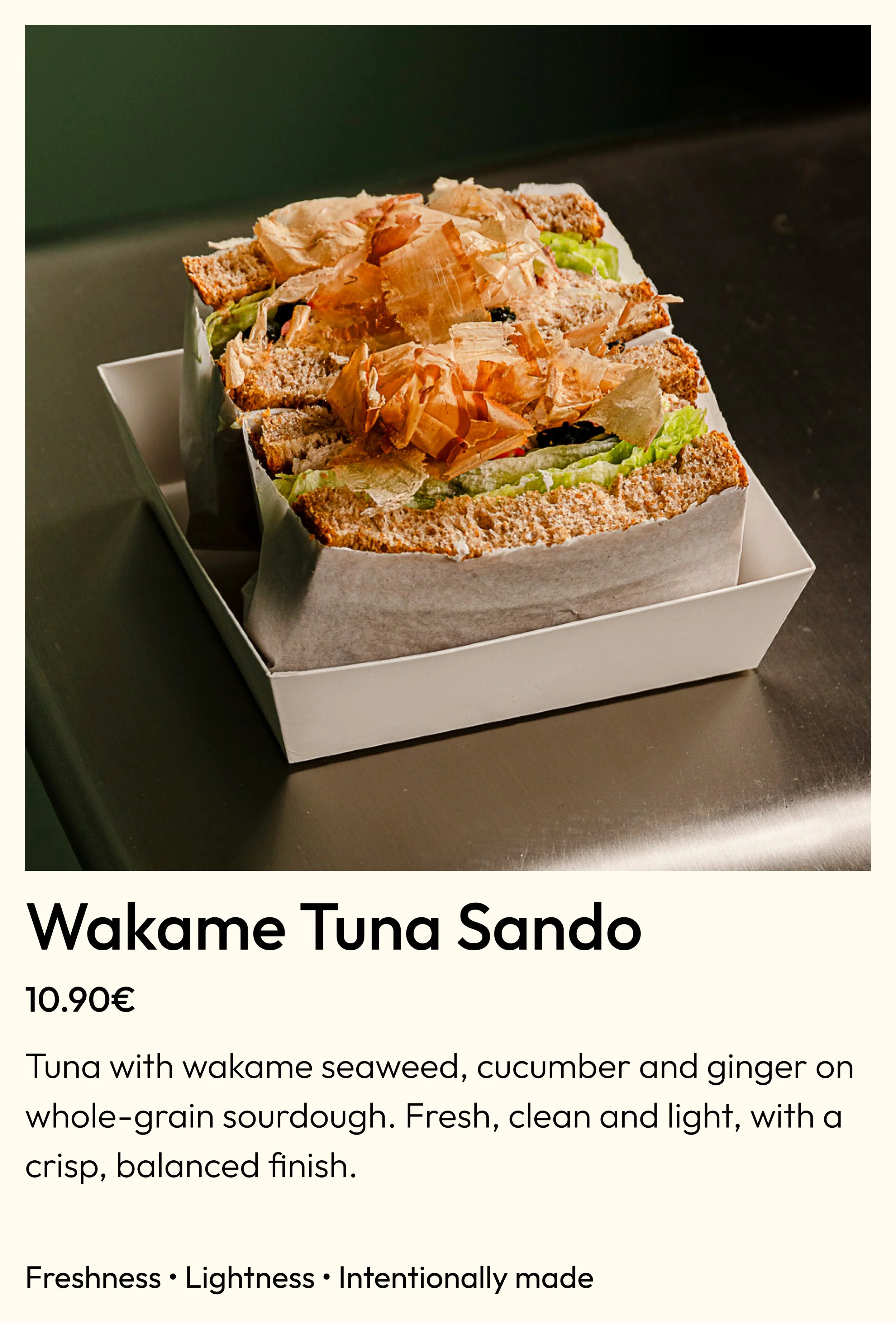 A whole grain sourdough sandwich with tuna, wakame seaweed, cucumber, and ginger, topped with bonito flakes and served in a white paper-lined box.