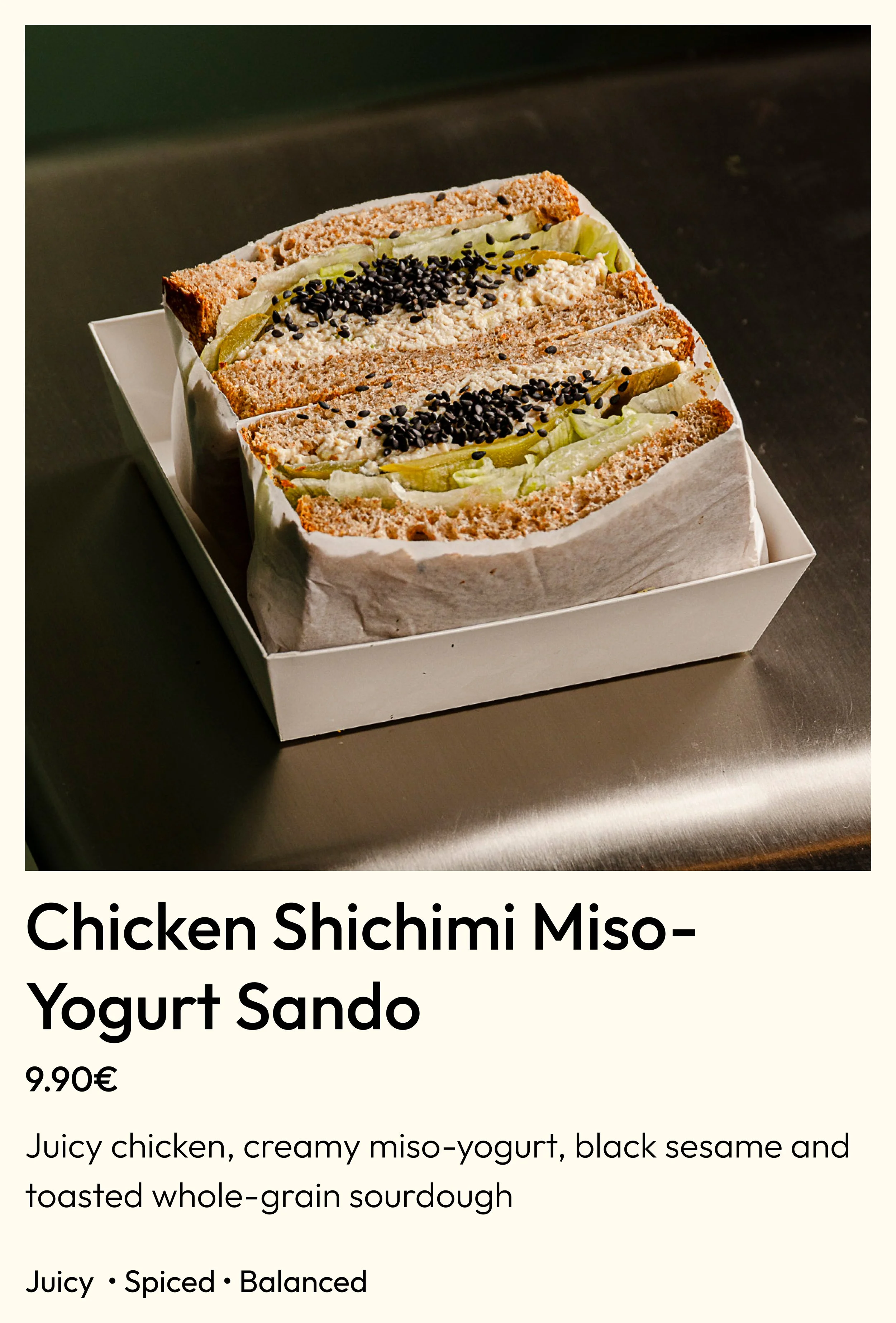 A sandwich cut into halves showing chicken, creamy miso-yogurt spread, black sesame seeds, and lettuce inside toasted whole-grain sourdough bread, placed in a white box.