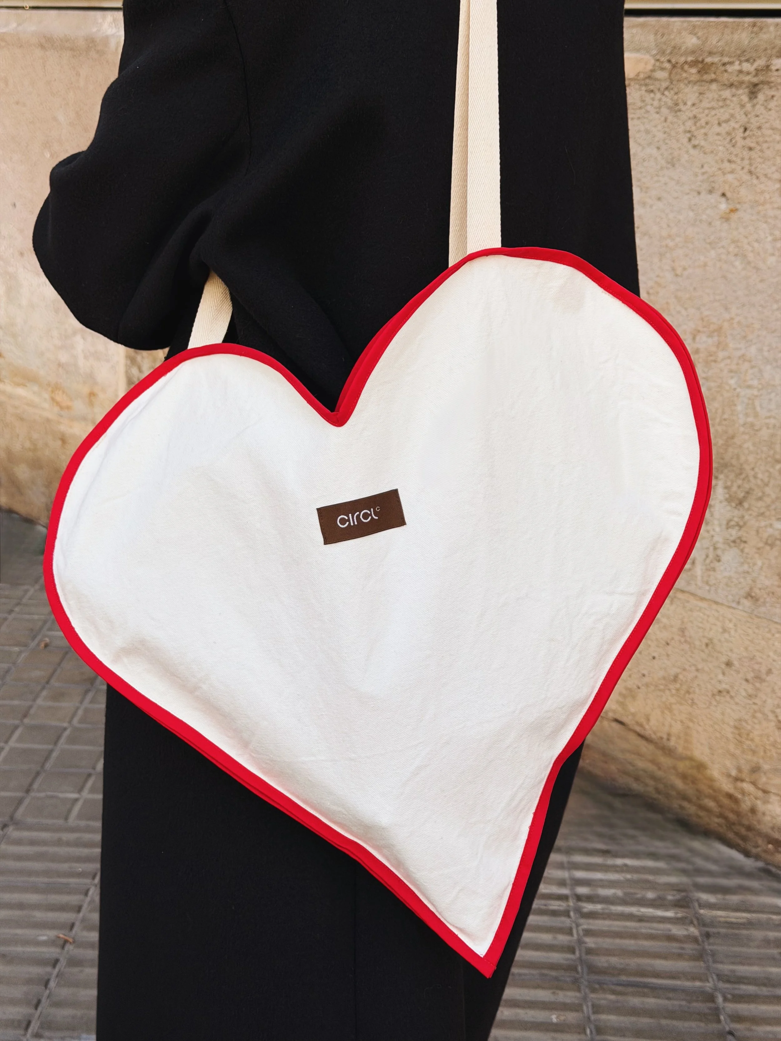 Person carrying a white heart-shaped bag with red piping and a black label that says 'CIRCL' on a shoulder strap.