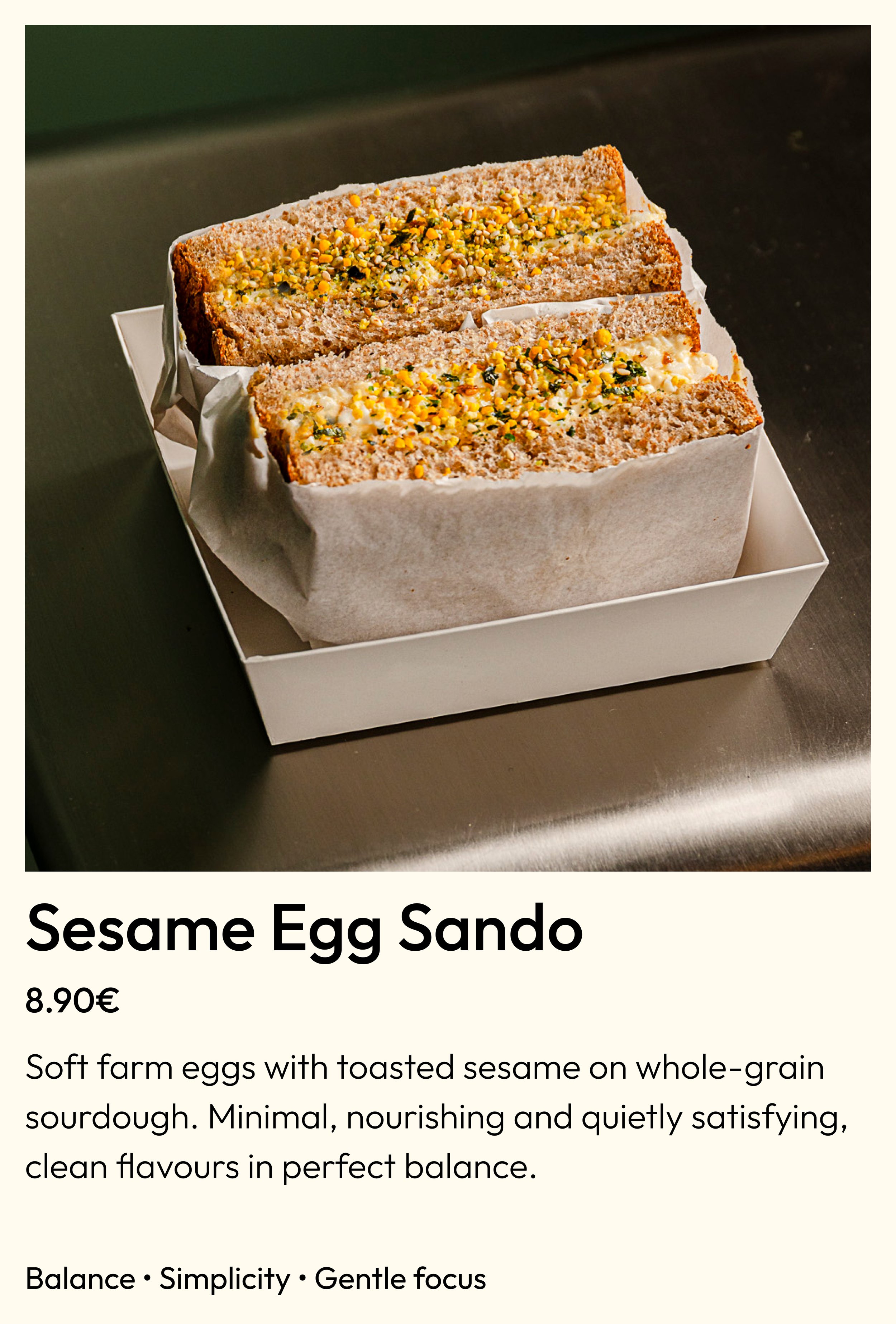 Close-up of two slices of sesame egg sando sandwiches with toasted sesame seeds, placed in a white paper-lined box on a dark surface.