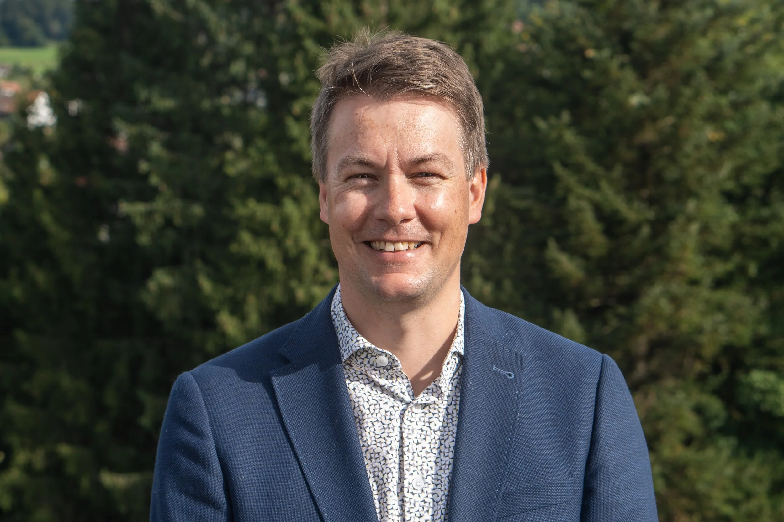 A man smiling outdoors, wearing a navy blazer and a patterned shirt, with trees in the background.