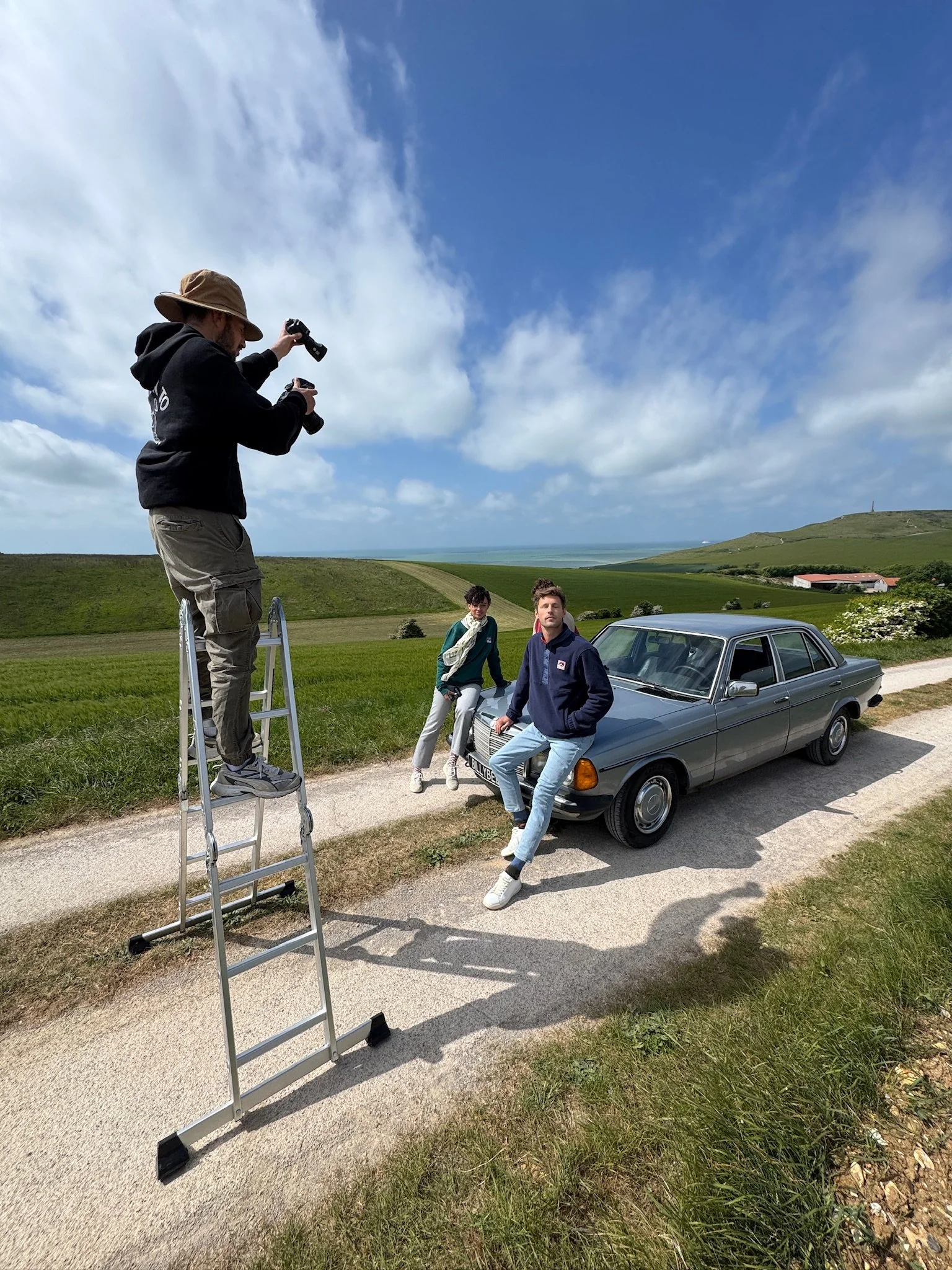 Deux jeunes adultes posent près d'une voiture vintage, en plein air avec un paysage de champs verts, un ciel partiellement nuageux et une horizon lointaine. Un photographe se tient sur une échelle pour prendre la photo.