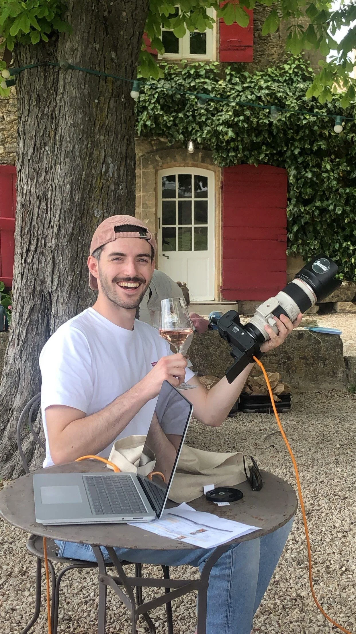 Un homme souriant, portant une casquette rose, tient un verre de rosé dans une main et un grand appareil photo dans l’autre, assis à une table en extérieur avec un ordinateur portable et des documents, devant un arbre avec une maison en pierre en arr