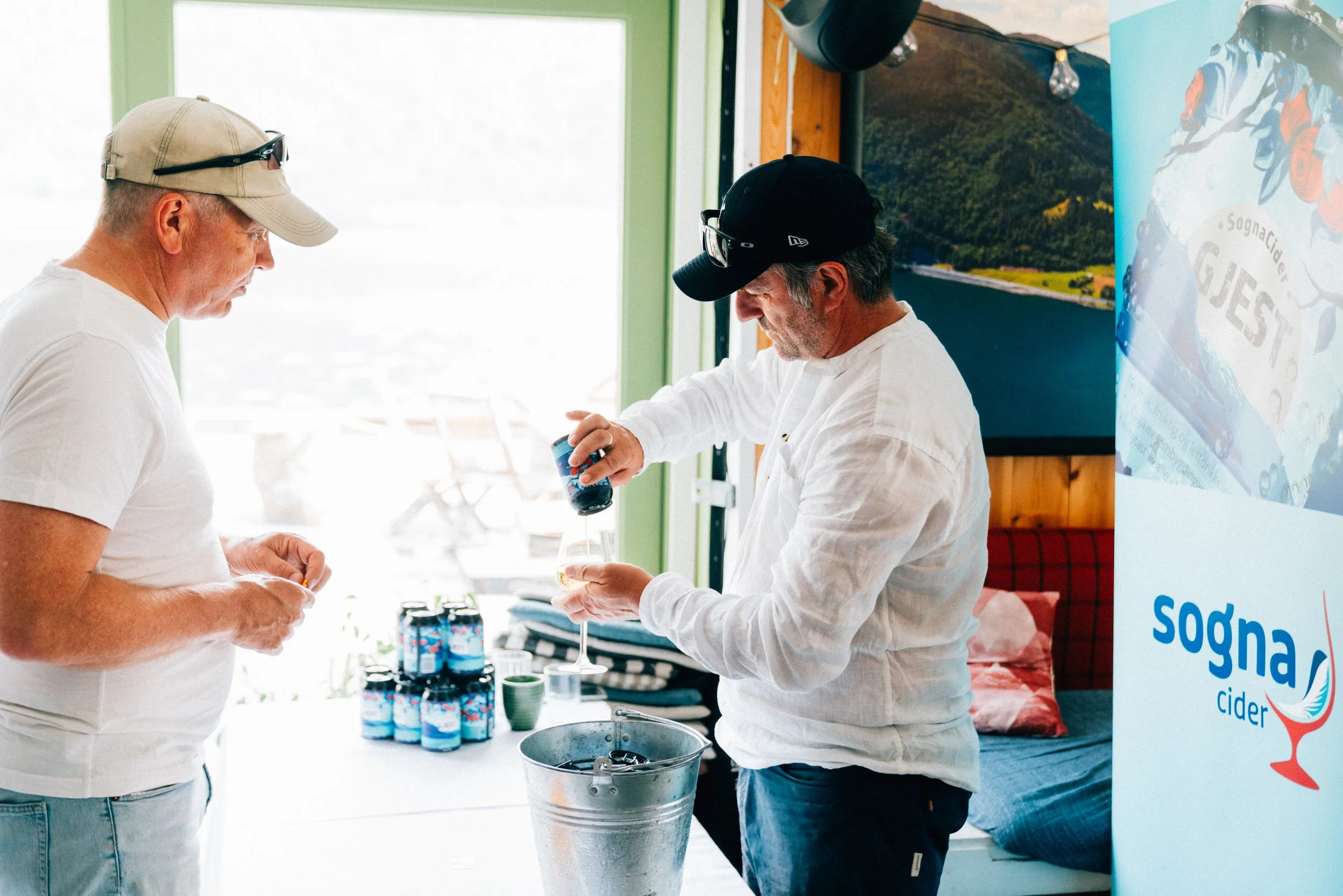 To menn står ved et bord og drikker sider, en av dem heller opp i et glass mens den andre ser på. Det er pakker med sider og en bordplate synlig, med vindu bak som slipper inn mye lys. En vertikal banner med logo og tekst til høyre.