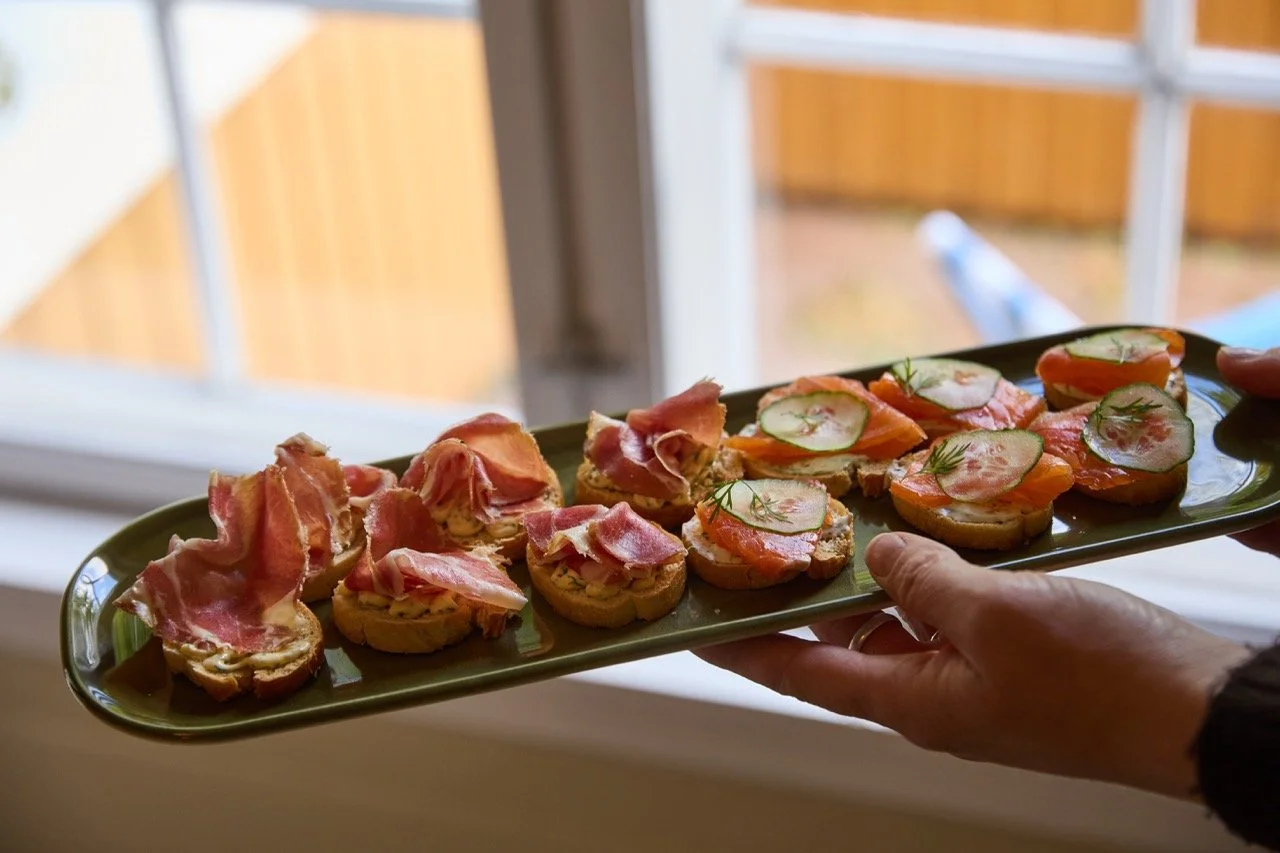 En person holder en lang, grønn serveringsfat med små blings til tapasmåltid, toppet med skinke, tomat og agurk.