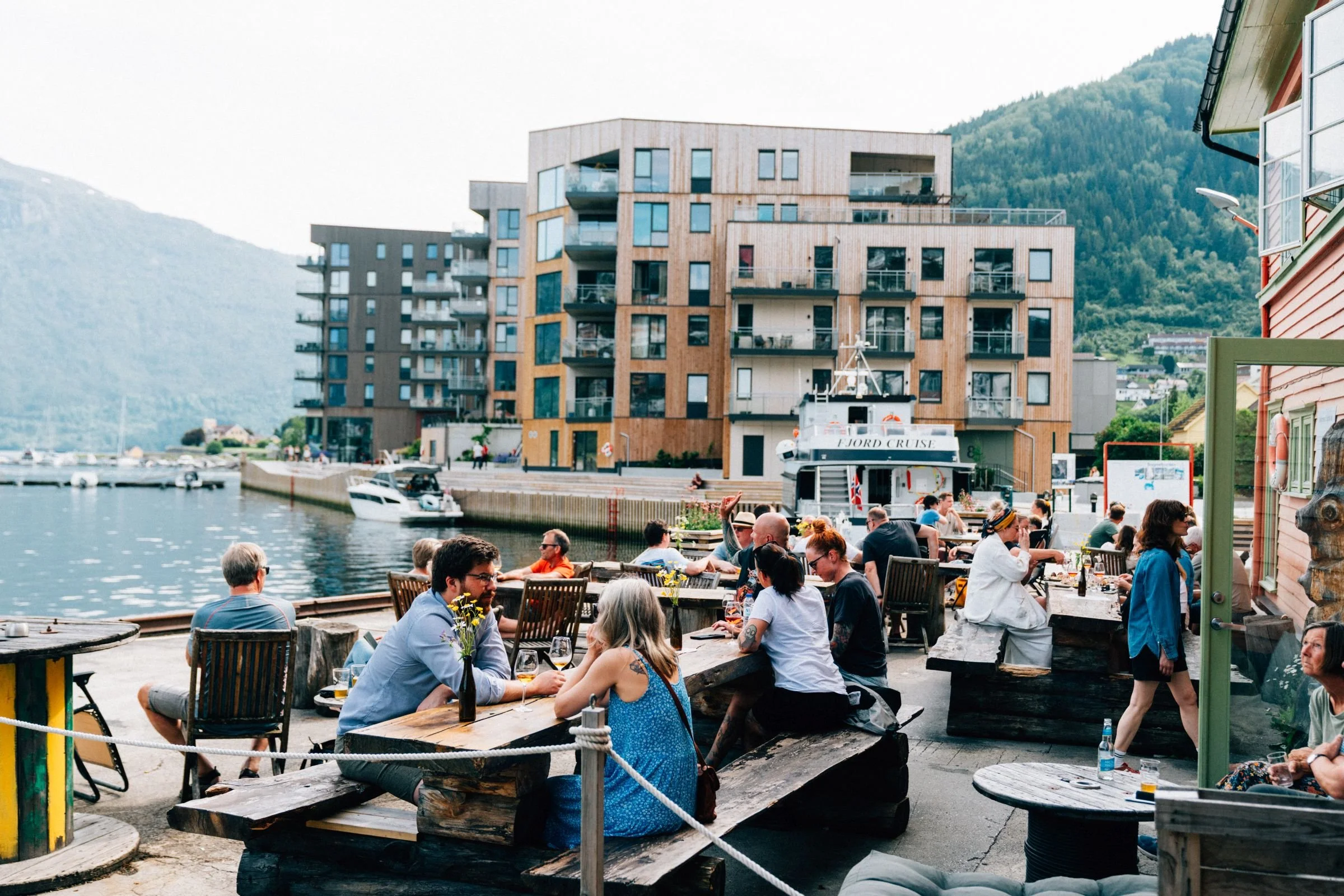 Mennesker spiser og henger ut ved en lokal pub eller restaurant ved vannet med moderne bygninger og fjell i bakgrunnen.