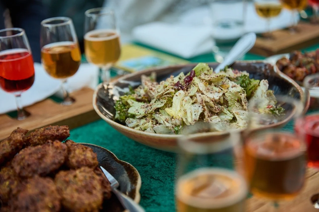 Bildet viser en biffesalat i en stor bolle, omgitt av glass med forskjellige fargede drikker, samt stekte kyllingvinger i forgrunnen. Bordet er dekket med grønne og hvite duker.