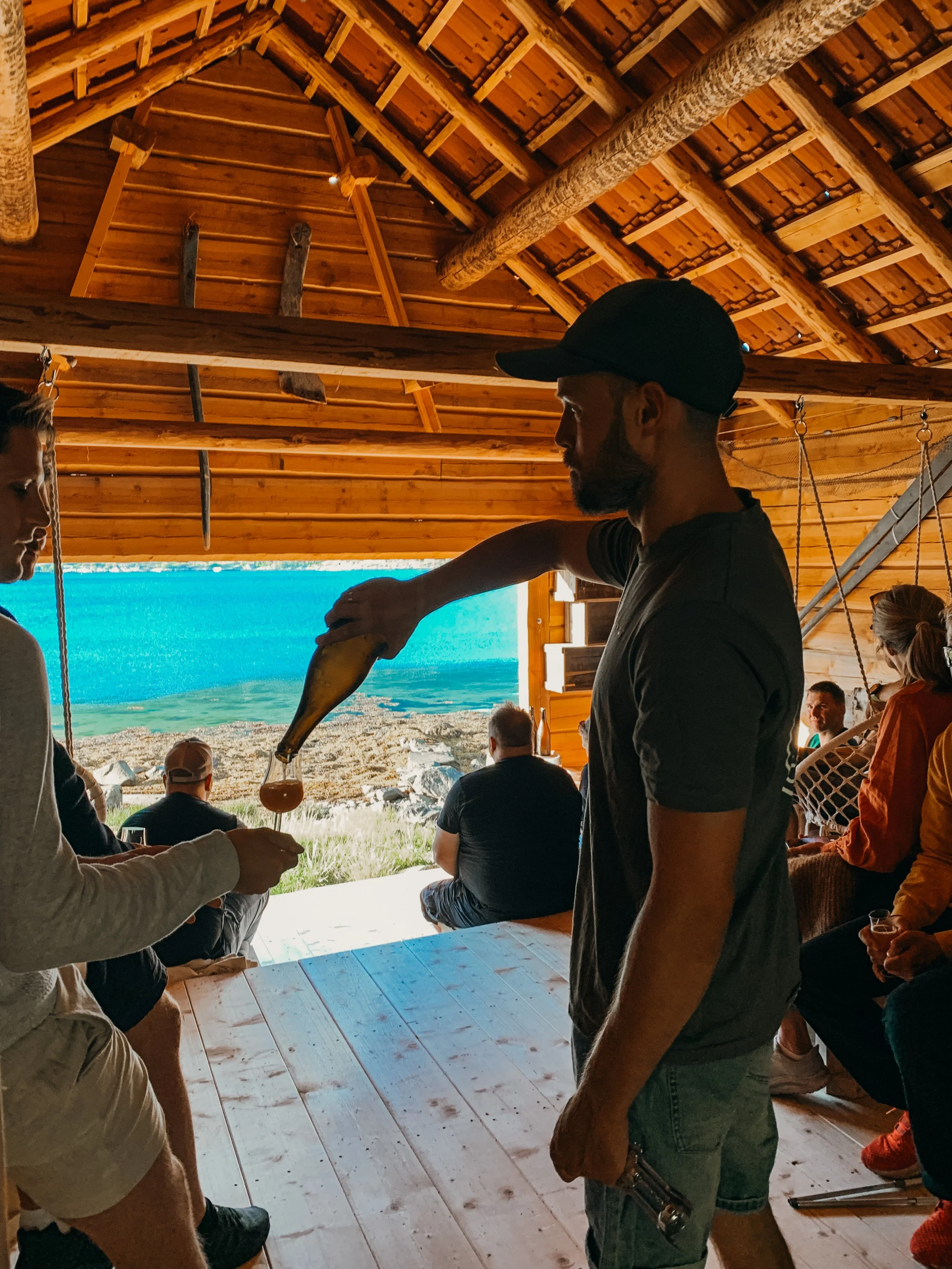 En mann i svart t-skjorte og Capri-shorts serverer drikke til en annen person i en trehytte med åpen utsikt over fjell og sjø.
