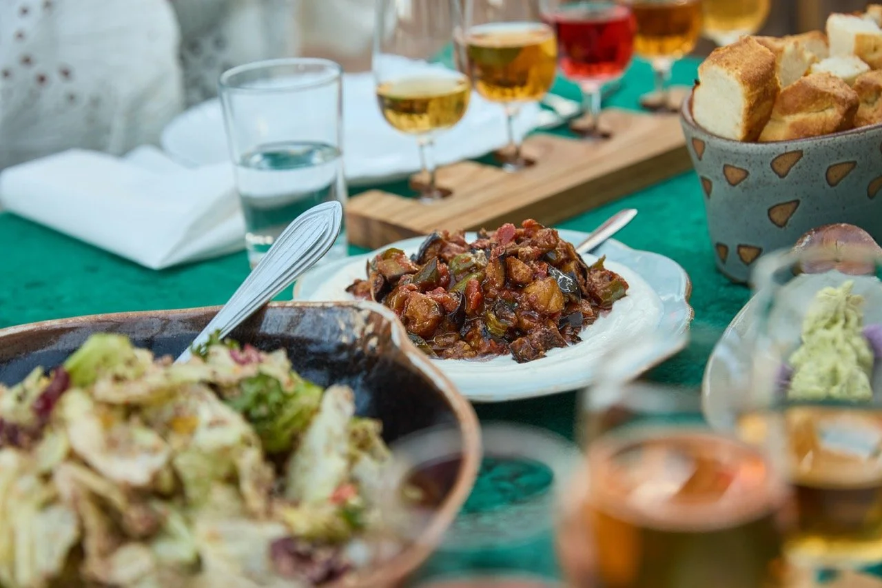 En bord fylt med ulike retter, inkludert en gryte med kjøtt- og grønnsaksblanding, en tallerken med gryte, flere glass med øl, og en skål med brød, på en grønn duk.