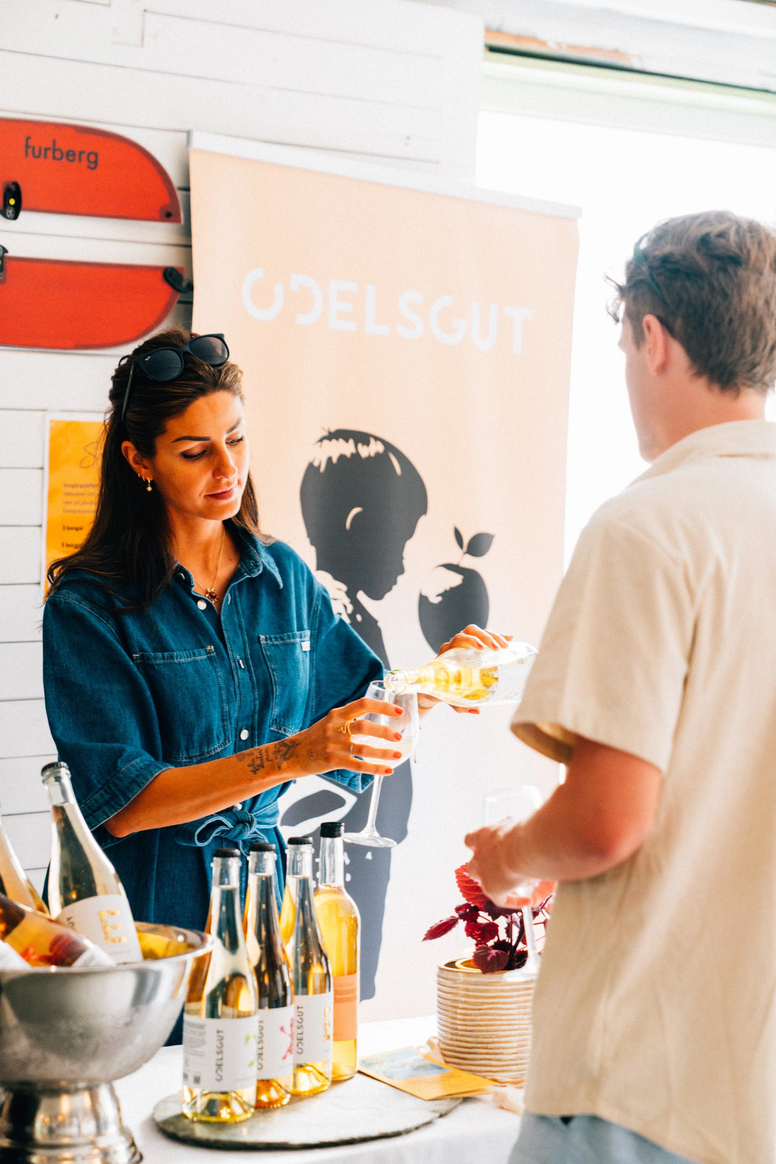 En kvinne i blå skjorte serverer hvitvin til en mann i beige skjorte ved en bod med flere vinflasker, en blomsterpotte og en logo med teksten 'ØDELSGUT'