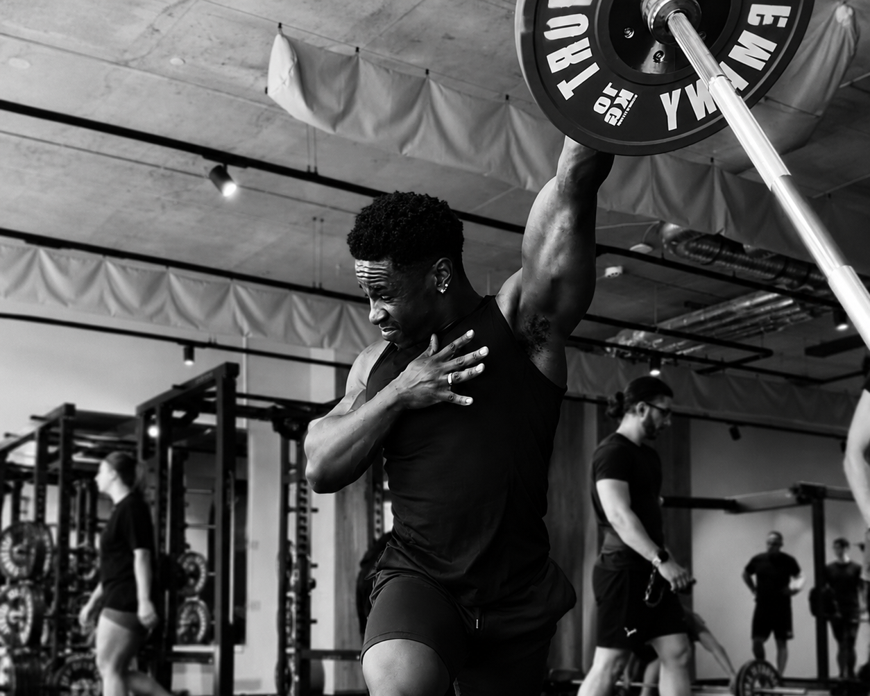 A man performing a weightlifting exercise in a gym, holding a barbell overhead with a facial expression of effort and focus.