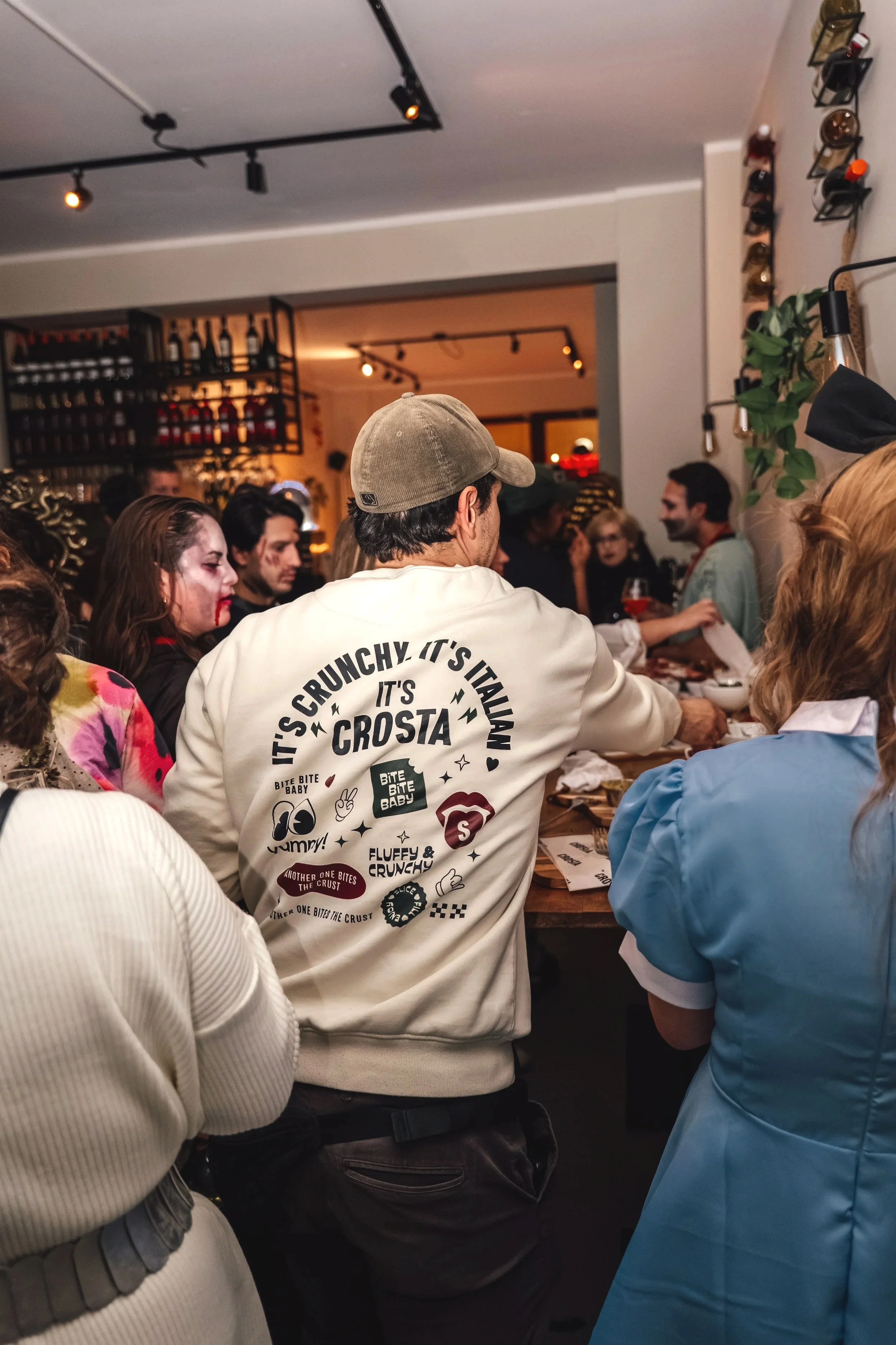 People gathered at a social event or party in a restaurant or bar, with one person in the center wearing a white jacket with playful graphics and text about crust and pizza, surrounded by others in costumes or themed clothing.