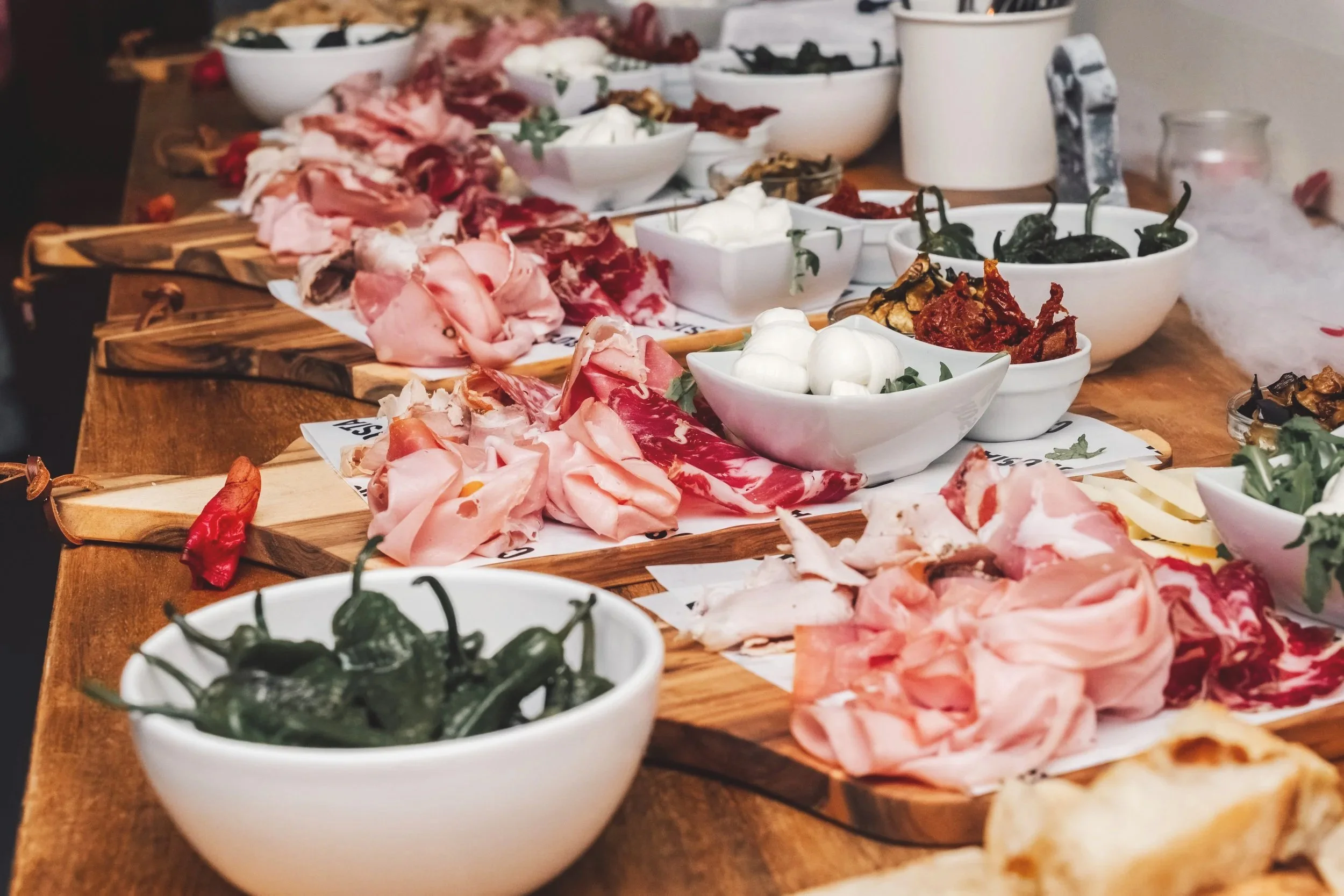 A variety of cold cuts and antipasti arranged on wooden serving boards and white bowls on a wooden table, including cured meats, cheese, peppers, and roasted vegetables.