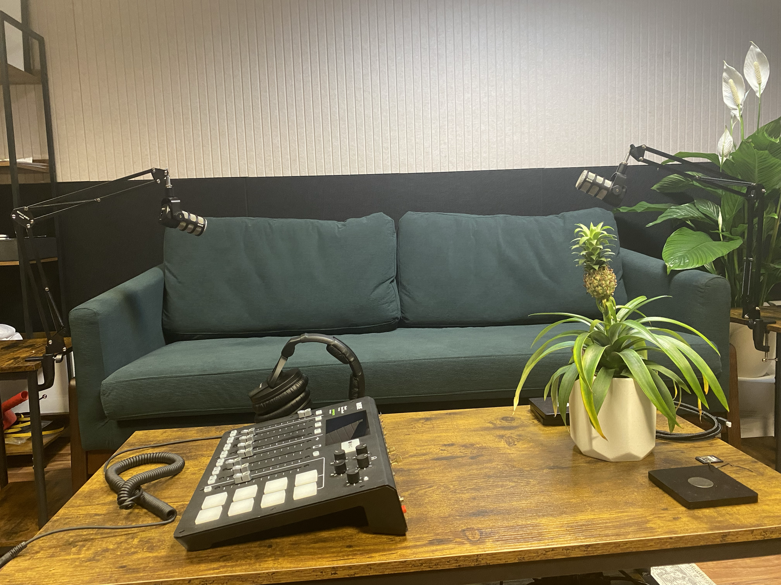 Podcast recording studio setup with a green sofa, two adjustable microphone arms, a sound mixer, a pair of headphones, a potted plant, and a black wireless charger on a wooden table.