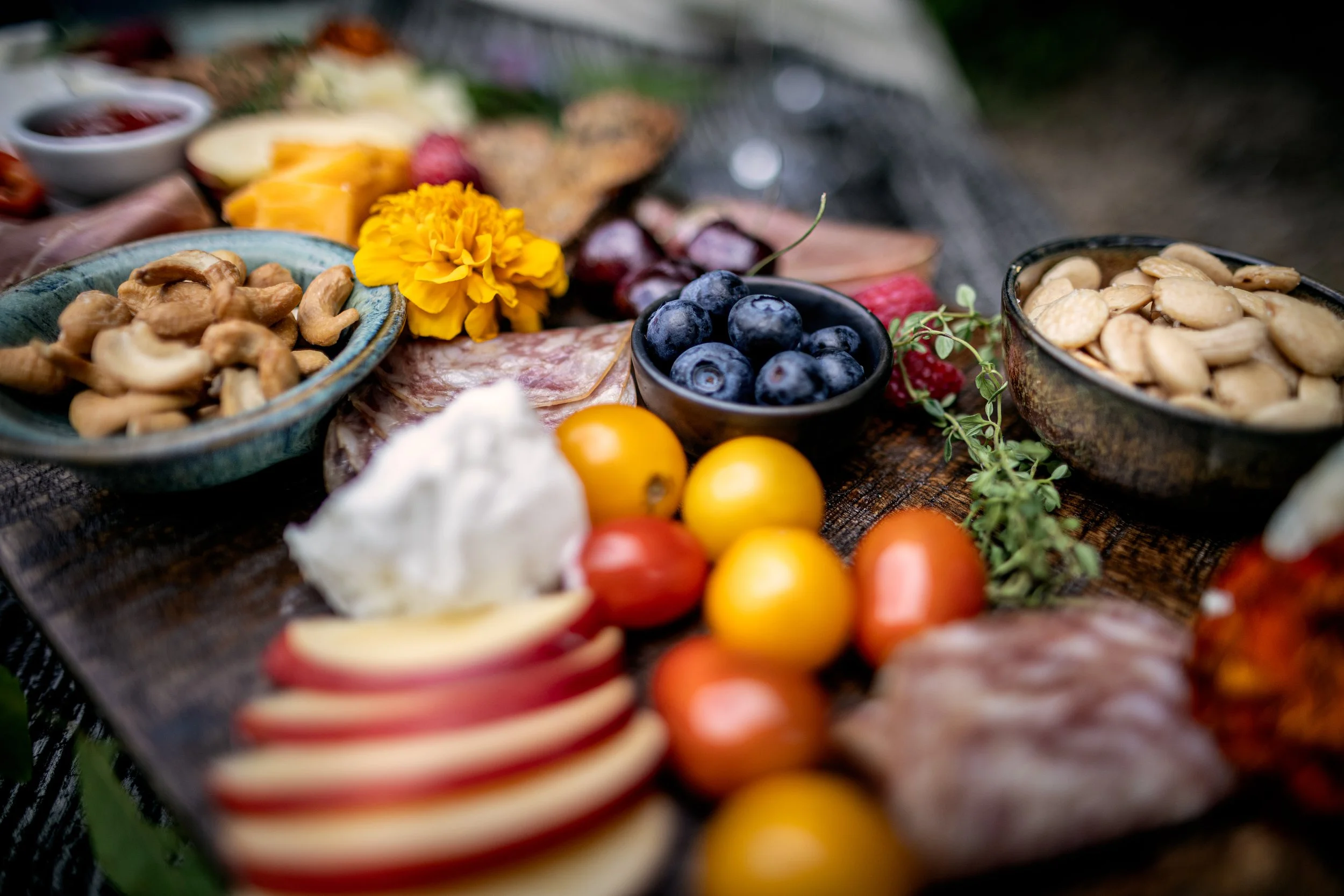 A cheese and charcuterie board with blueberries, cashews, tomatoes, apples, sliced cheeses, cured meats, and a yellow flower for decoration.