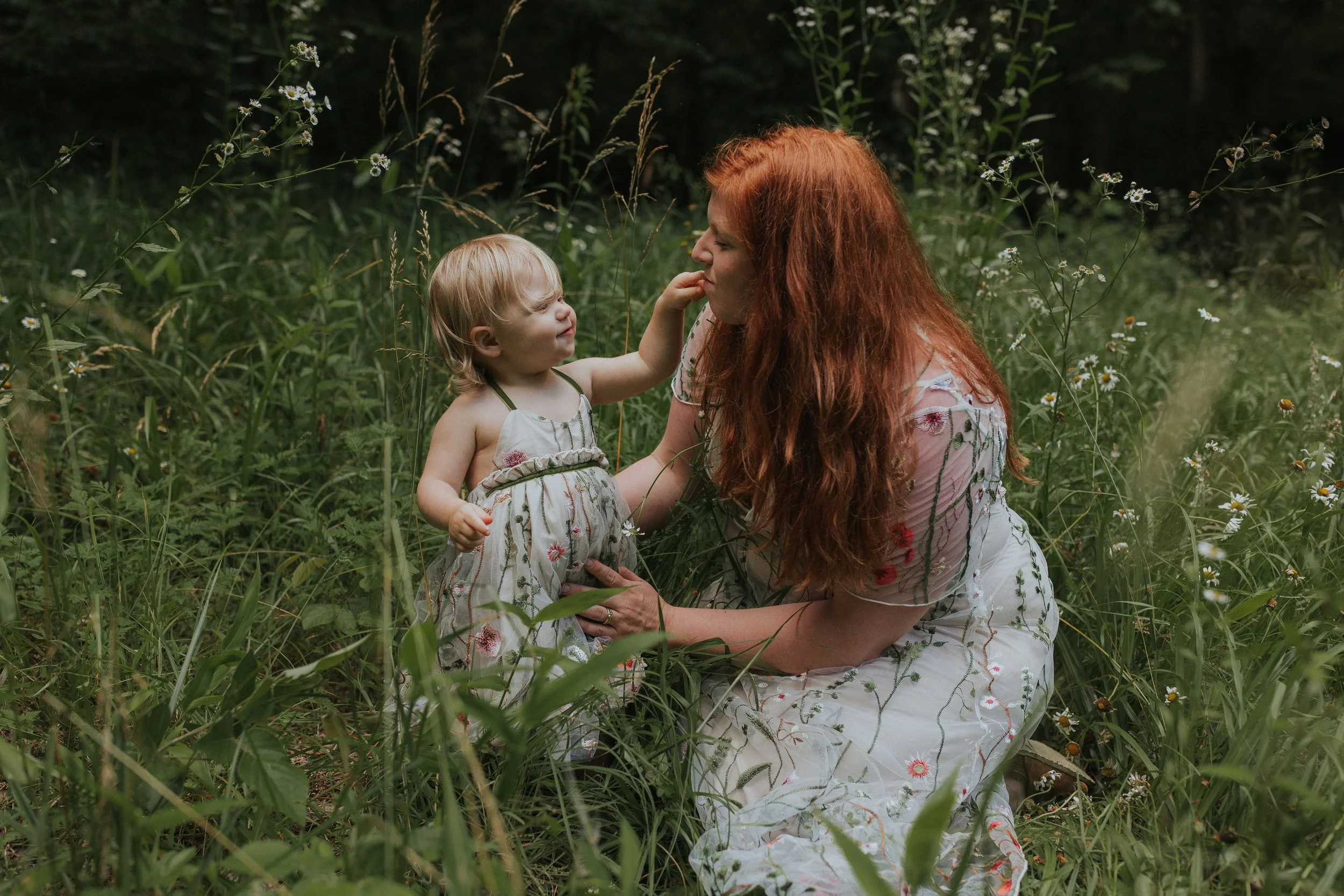 A red-haired woman and a young girl with blonde hair in a grassy field with flowers. The girl touches the woman's face.
