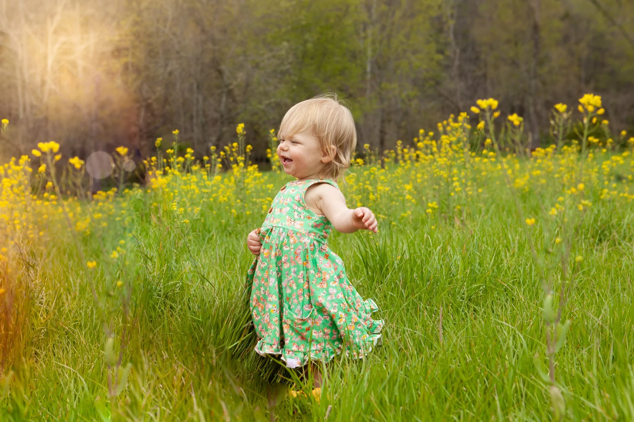 A young girl in a floral dress playing in a grassy field with yellow flowers and trees in the background on a sunny day.