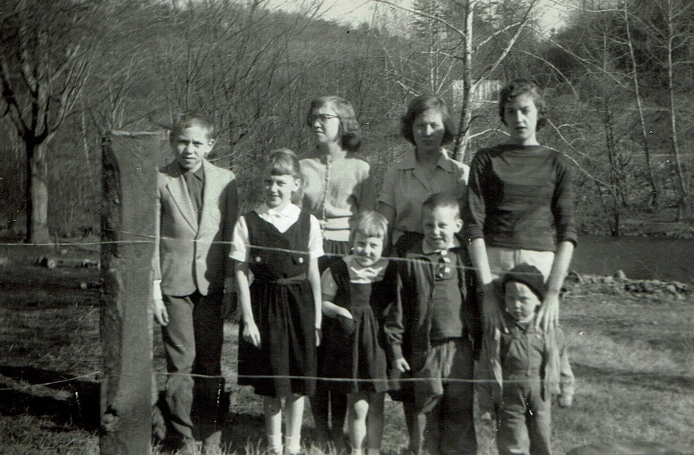 A black and white photo of a group of eight children standing outdoors by a wire fence, with trees and a body of water in the background, during daytime.