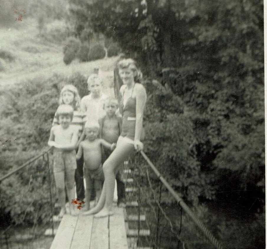 Black and white photo of seven people, including children and a woman, standing on a small wooden bridge with rope railings outdoors in a forested area.