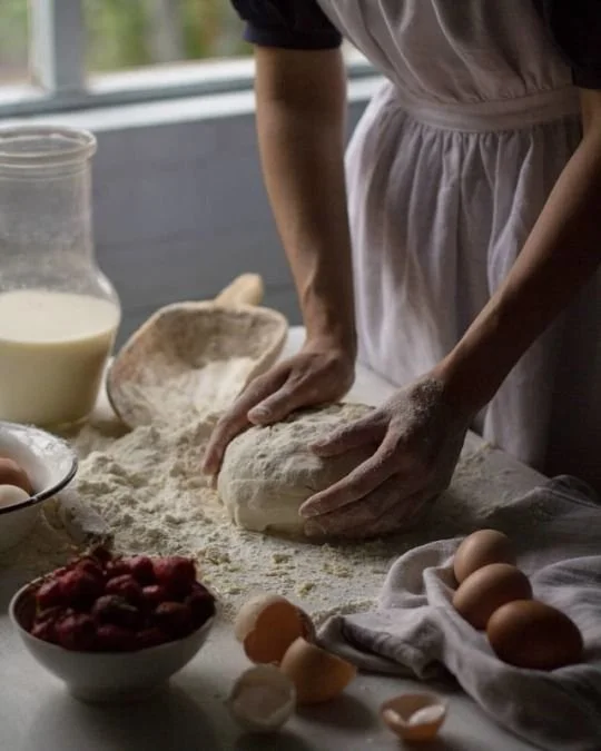 A person wearing a white apron is kneading dough on a floured surface with eggs, berries, and baking ingredients nearby.