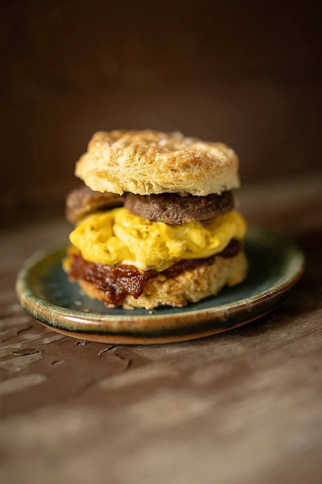 Close-up of a breakfast sandwich with an English muffin, scrambled eggs, bacon, sausage patty, and a biscuit, served on a small green plate on a rustic wooden surface.