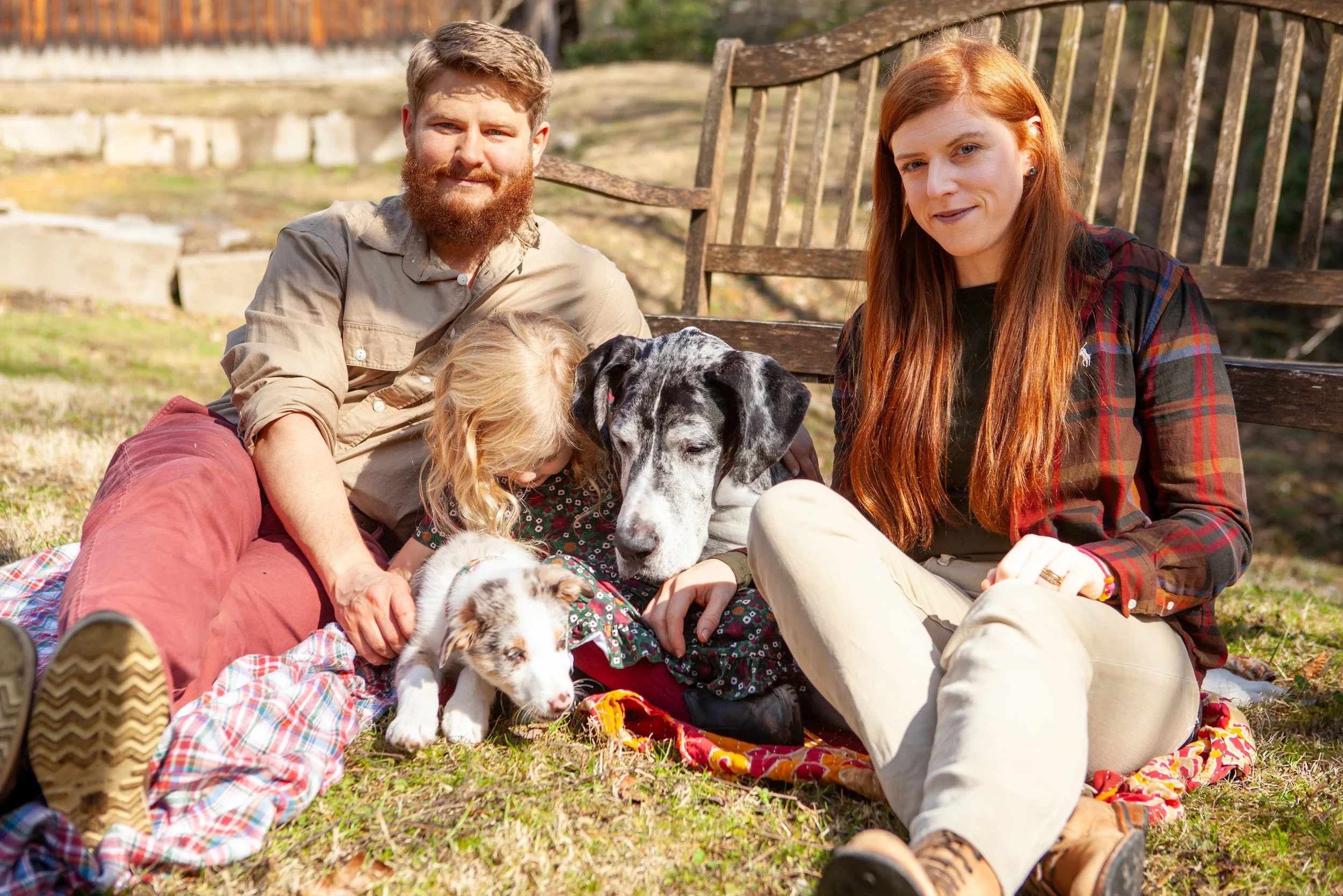 A family with two dogs sitting on the grass outdoors in front of a wooden fence, enjoying a sunny day.