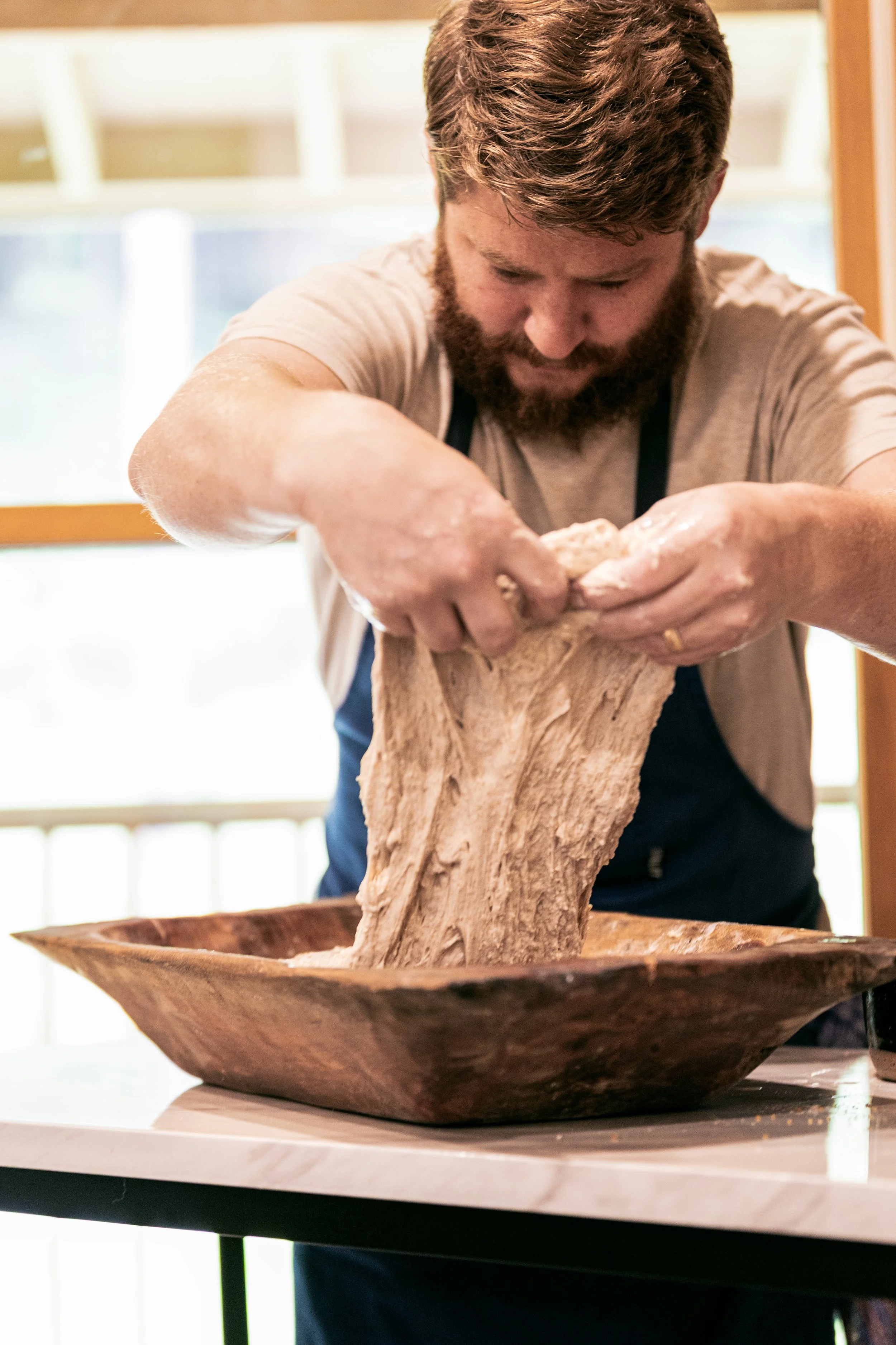 A man with a beard and short hair kneels over a wooden bowl, stretching and kneading dough.
