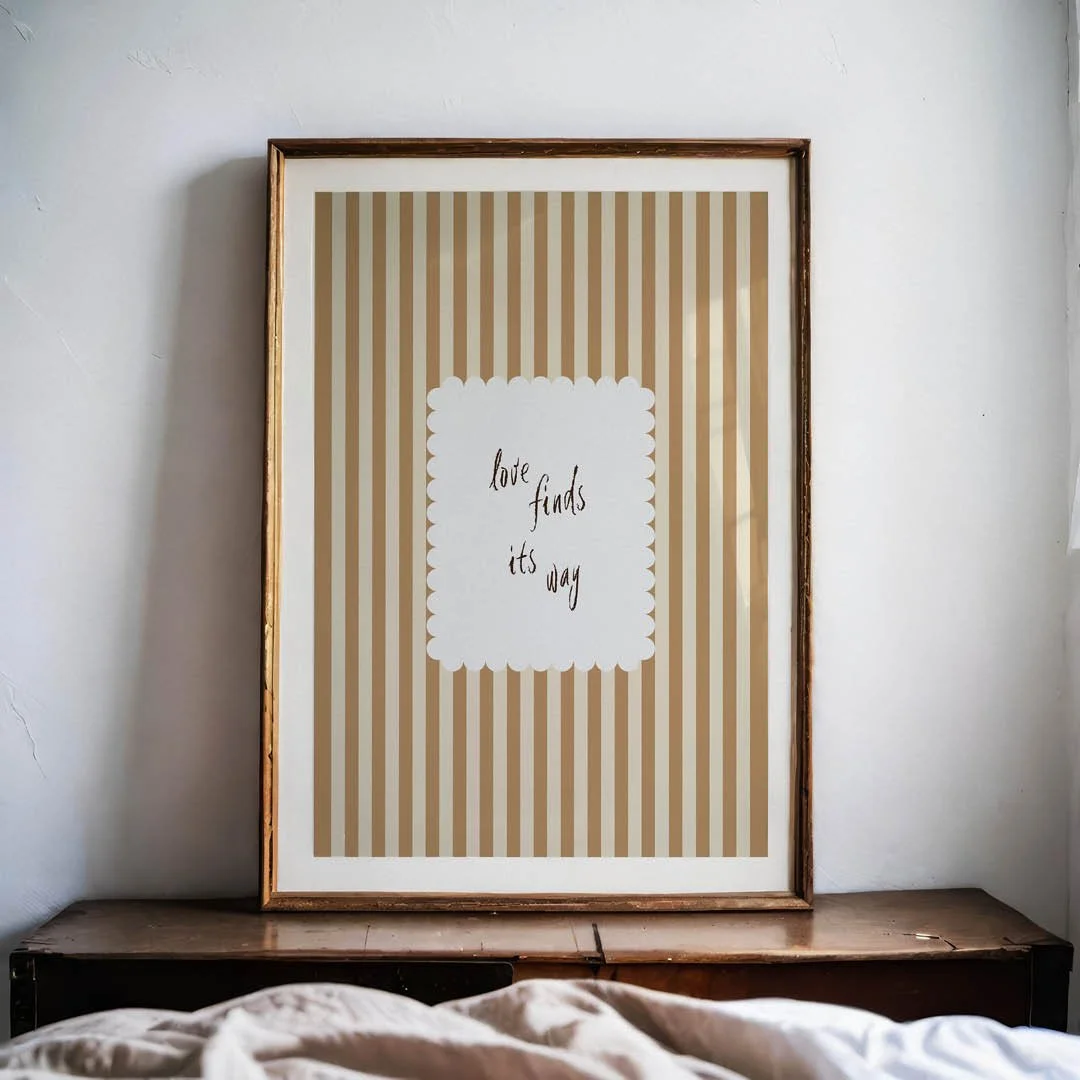 Framed poster featuring vertical tan and cream stripes, with a central scalloped white square containing the cursive phrase "love finds its way."
