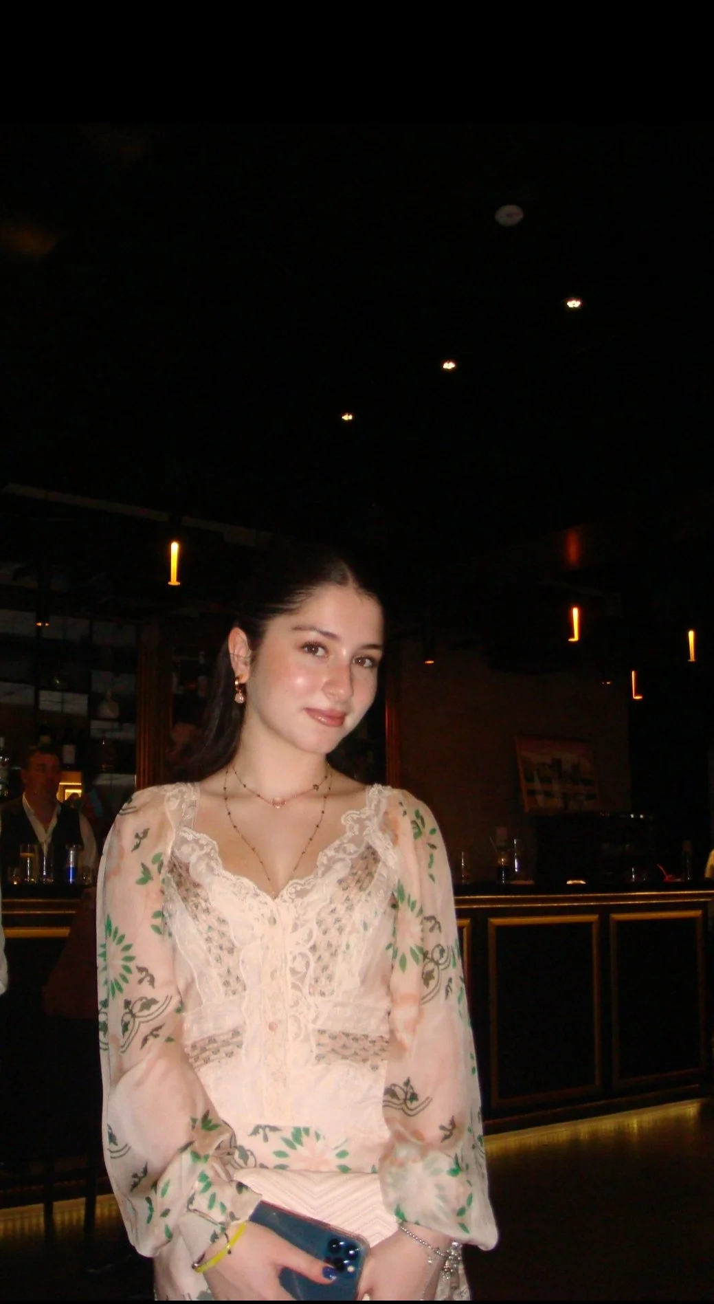 A young woman with dark hair dressed in a light-colored, embroidered dress, standing in a dimly lit room, possibly a restaurant or bar, holding a smartphone in her right hand.