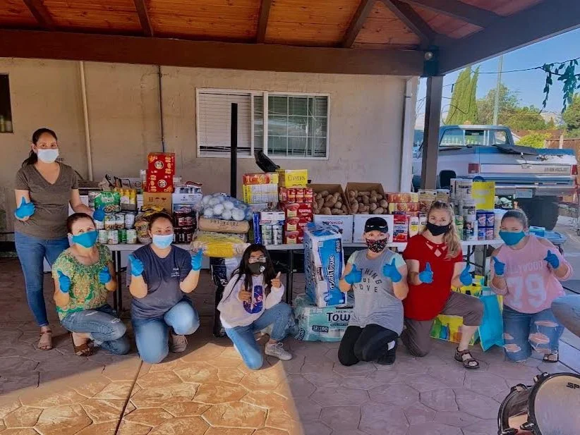 Group of people wearing face masks and gloves, giving thumbs up, standing behind a table filled with food donations outside a building.