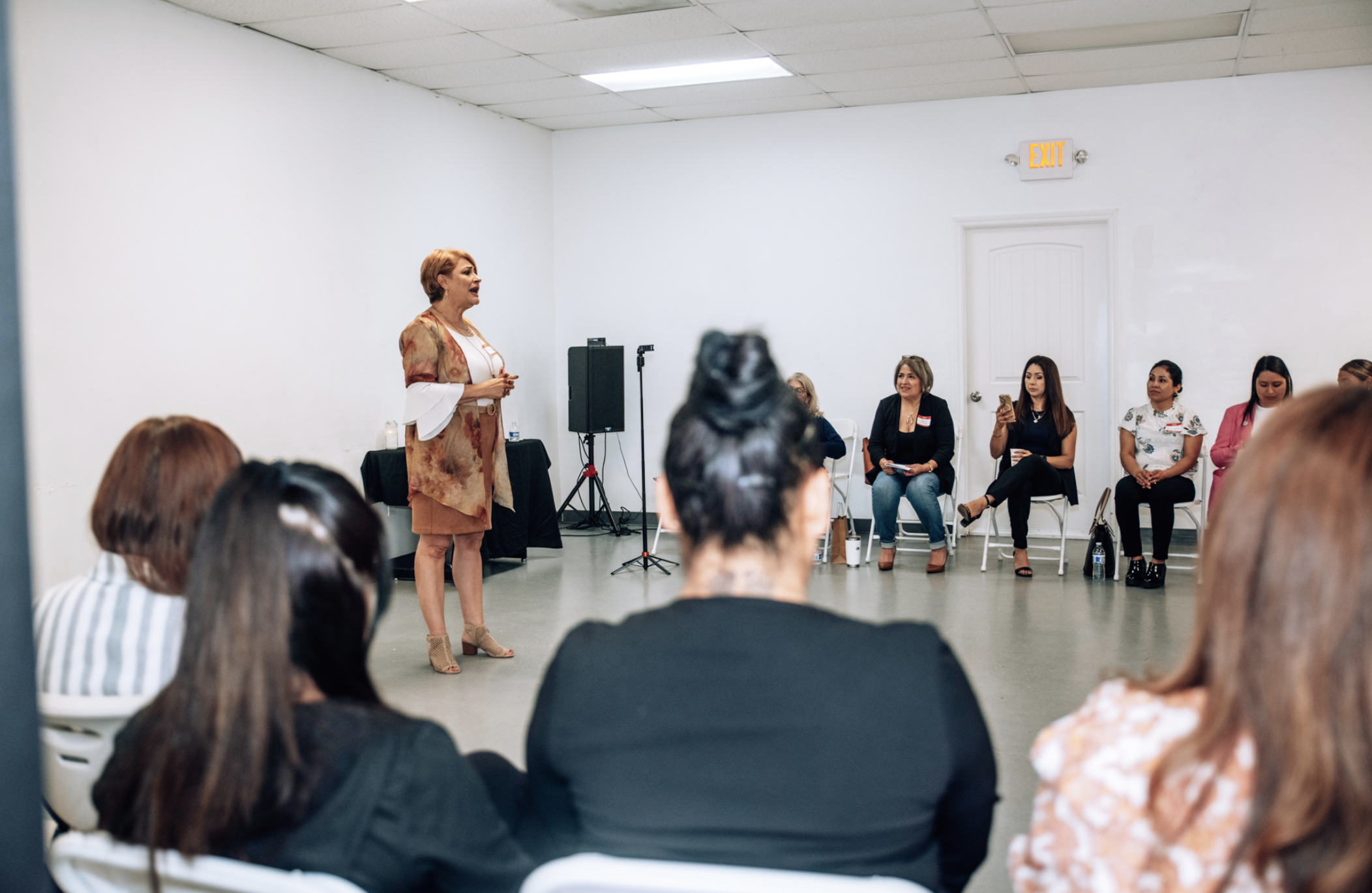A woman with short reddish hair speaking to an audience in a white-walled room. Several women are seated, some taking notes or photos, listening to her presentation.
