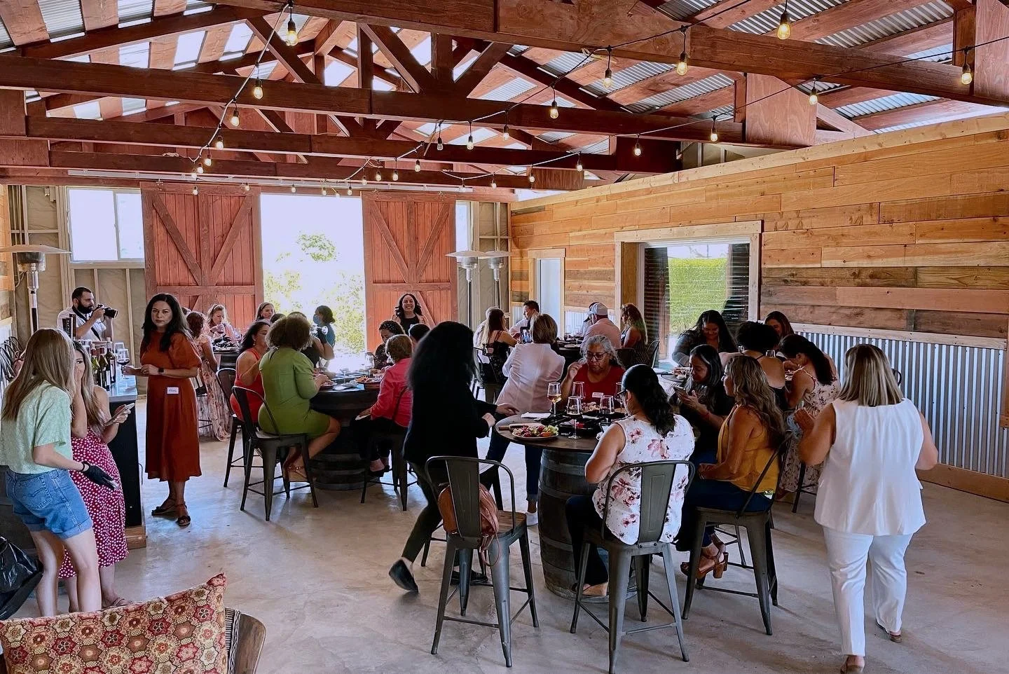 People gathering inside a rustic, wooden barn with string lights, sitting at tables and standing, during a social event or celebration.