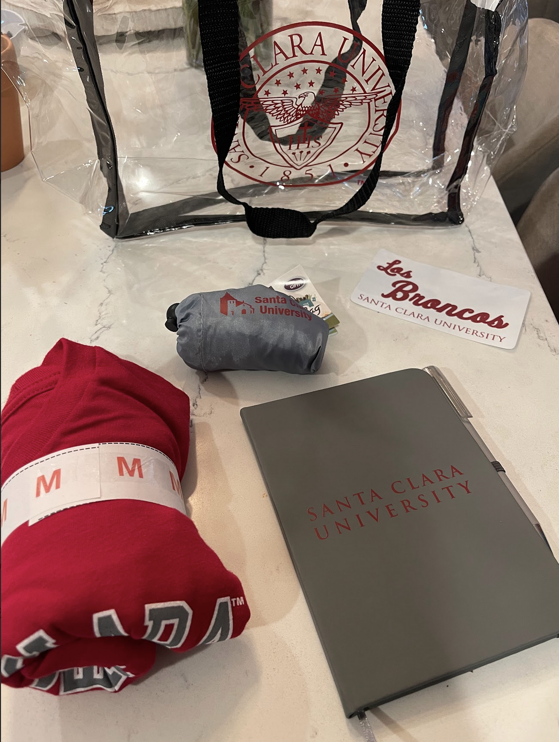Items related to Santa Clara University on a white marble table, including a clear bag with the university's logo, a red hoodie, a gray water bottle, a black notebook, a name tag that reads "Los Broncos," and a gray pouch.