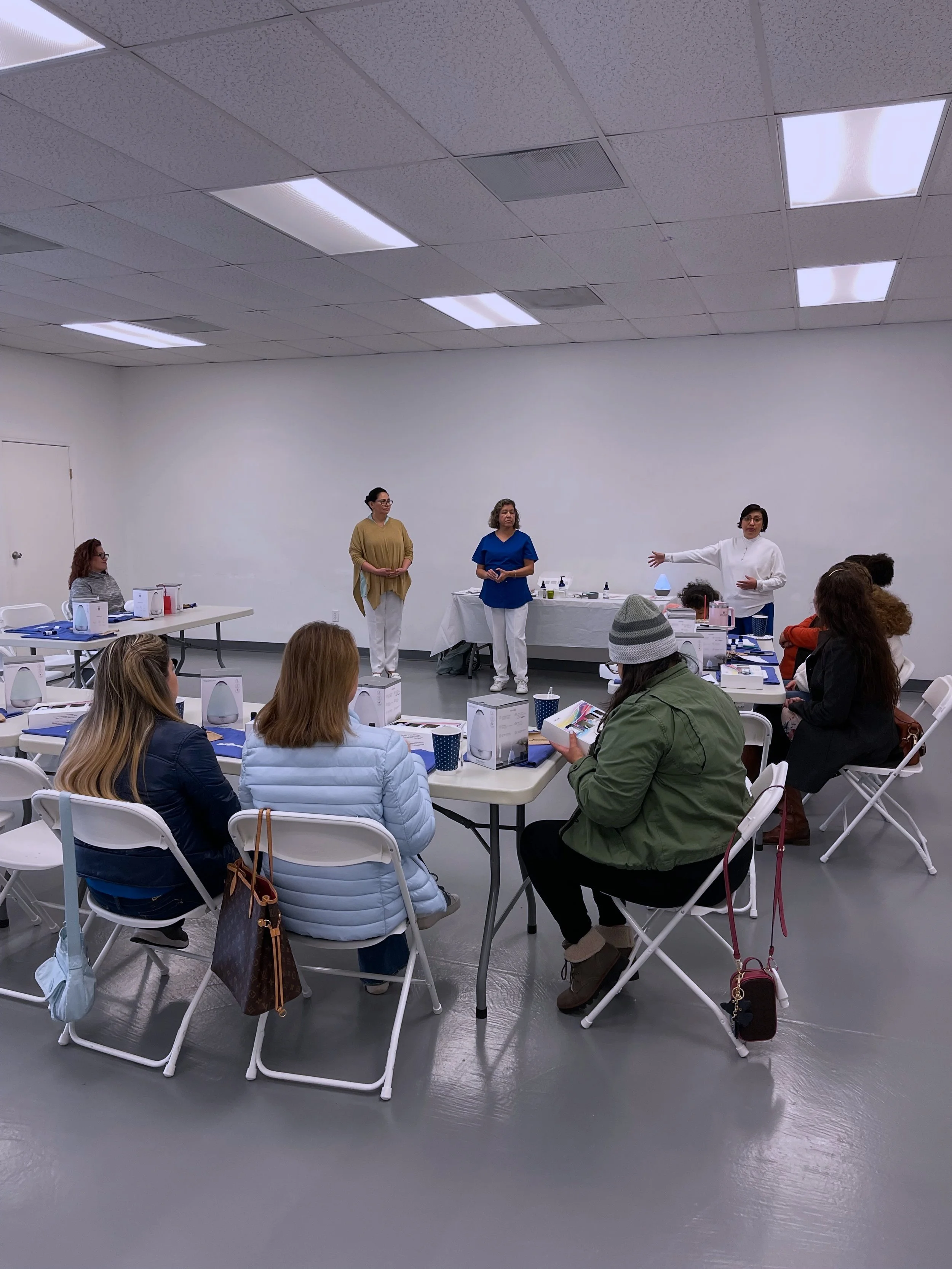 A group of women attending a workshop or class, seated at tables, listening to a speaker in white, with two other women standing nearby in a plain, white room.