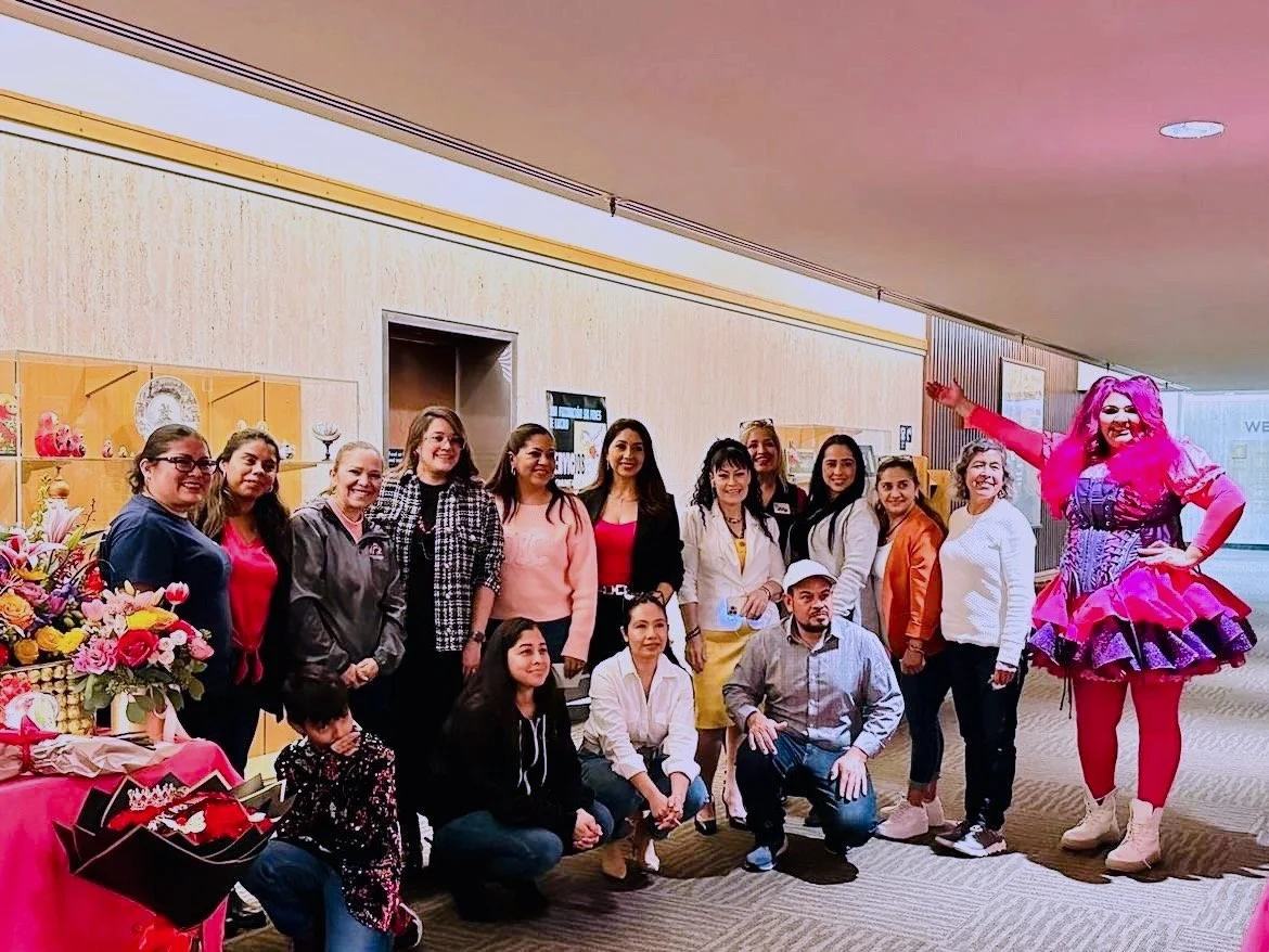 Group of people posing for a photo indoors, with a person dressed as a colorful clown or performer on the right side.