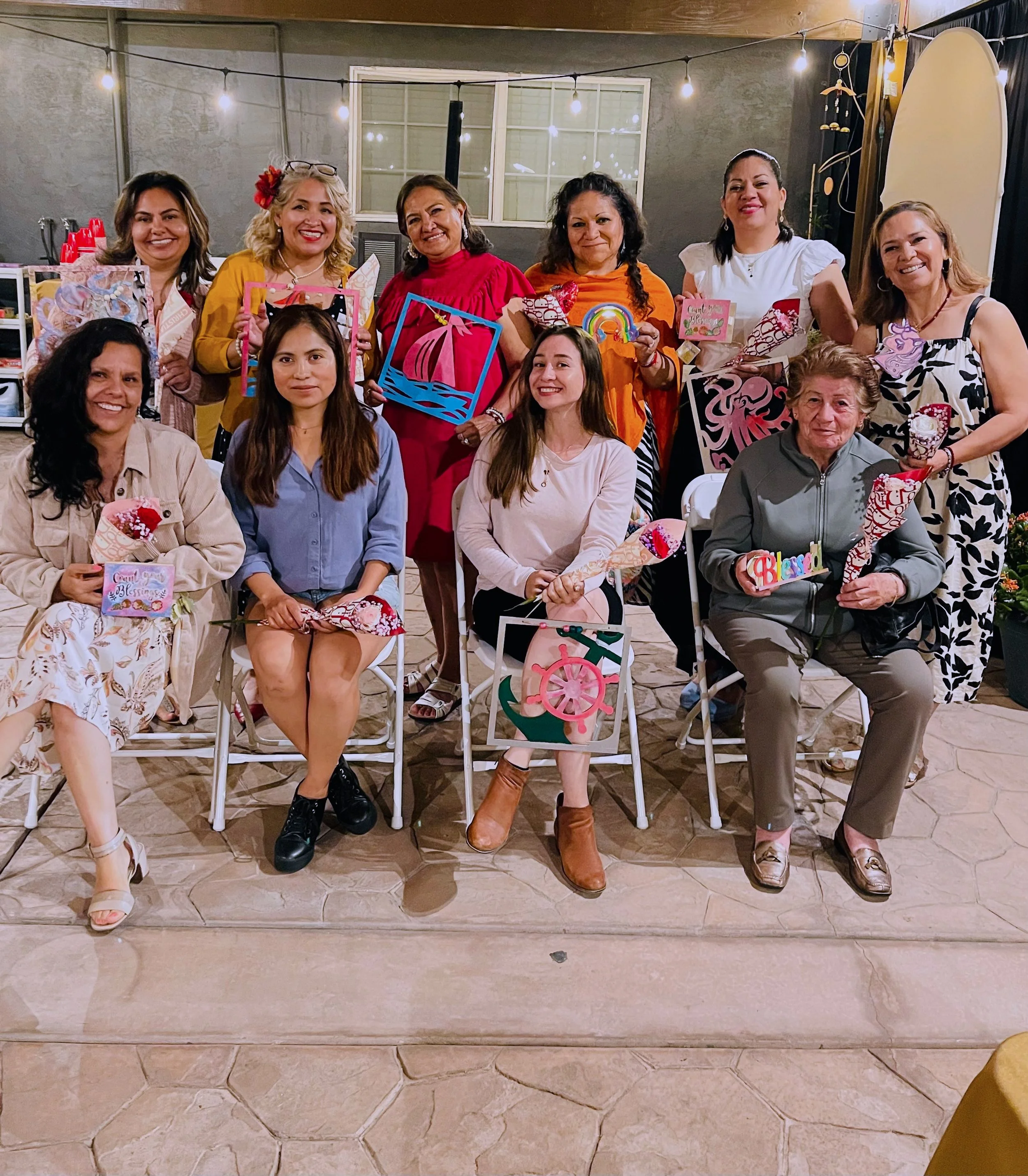 Group of women celebrating a birthday, holding decorated cookies and gifts, with a festive background decorated with string lights.