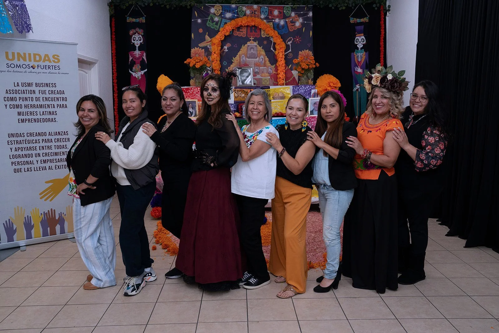 Group of ten women standing in front of a Dia de los Muertos altar, smiling and holding each other, dressed in casual and traditional outfits with colorful Día de los Muertos face paint and accessories.