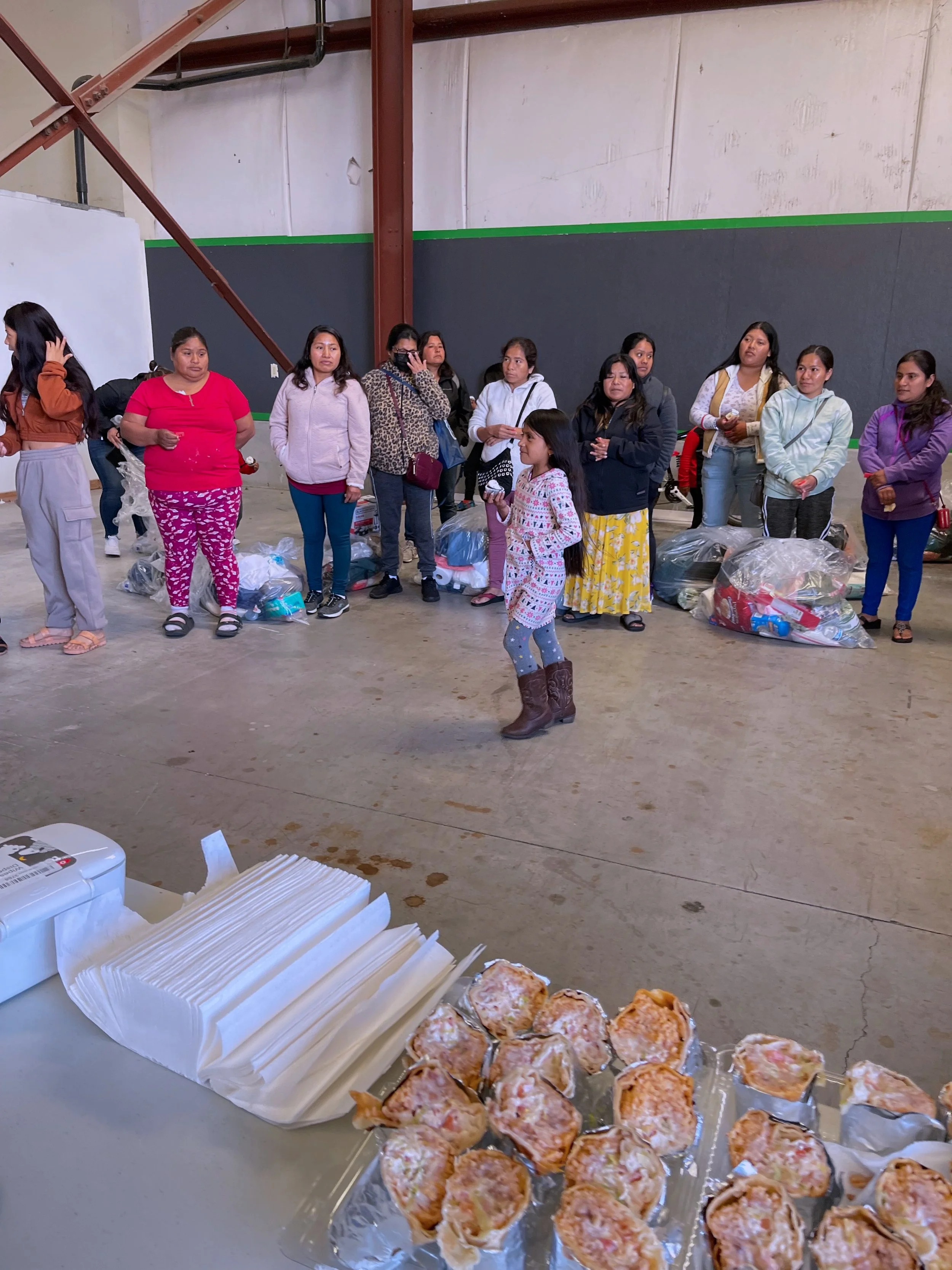A group of women and children standing inside a warehouse, with some sitting or kneeling on the floor, beside bags of supplies. In the foreground, a table holds napkins and plates of pizza pretzels.