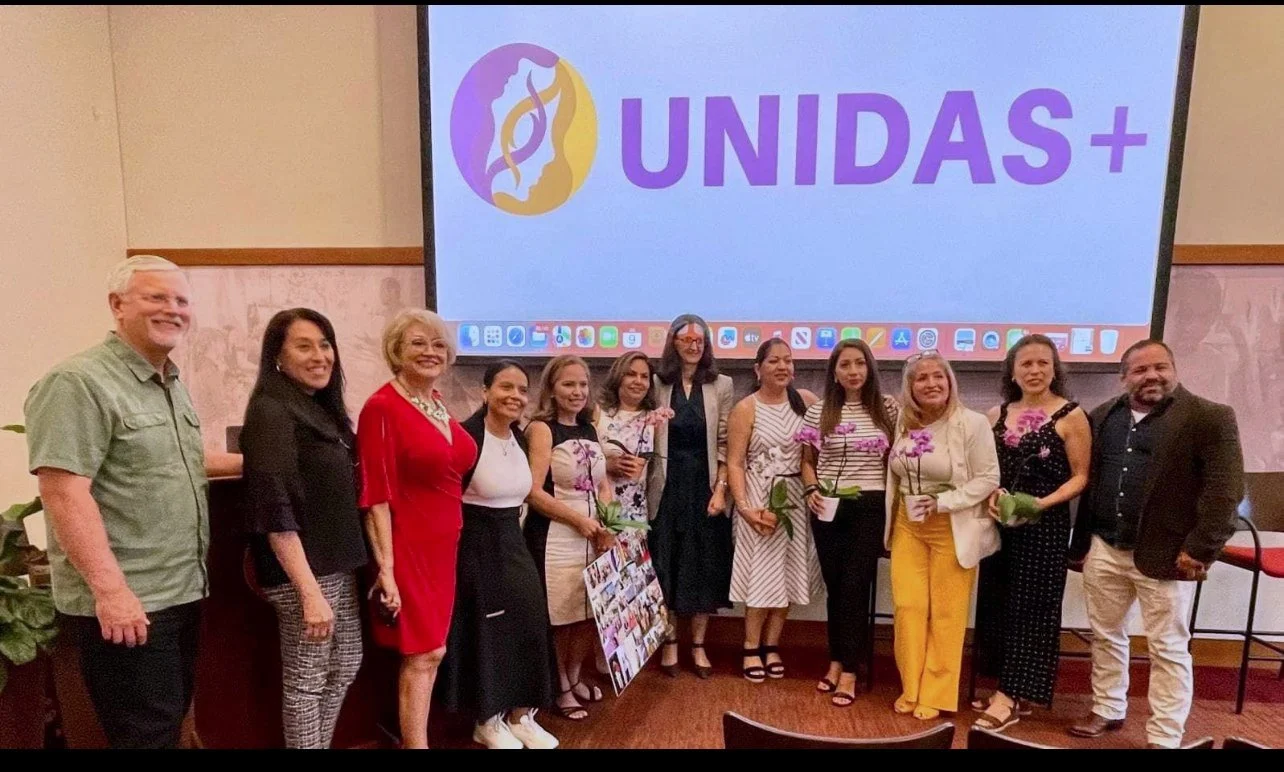 Group of eleven people standing in front of a large screen with the UNIDAS+ logo. The group includes men and women dressed in business casual and formal attire, holding flowers, at an indoor event.