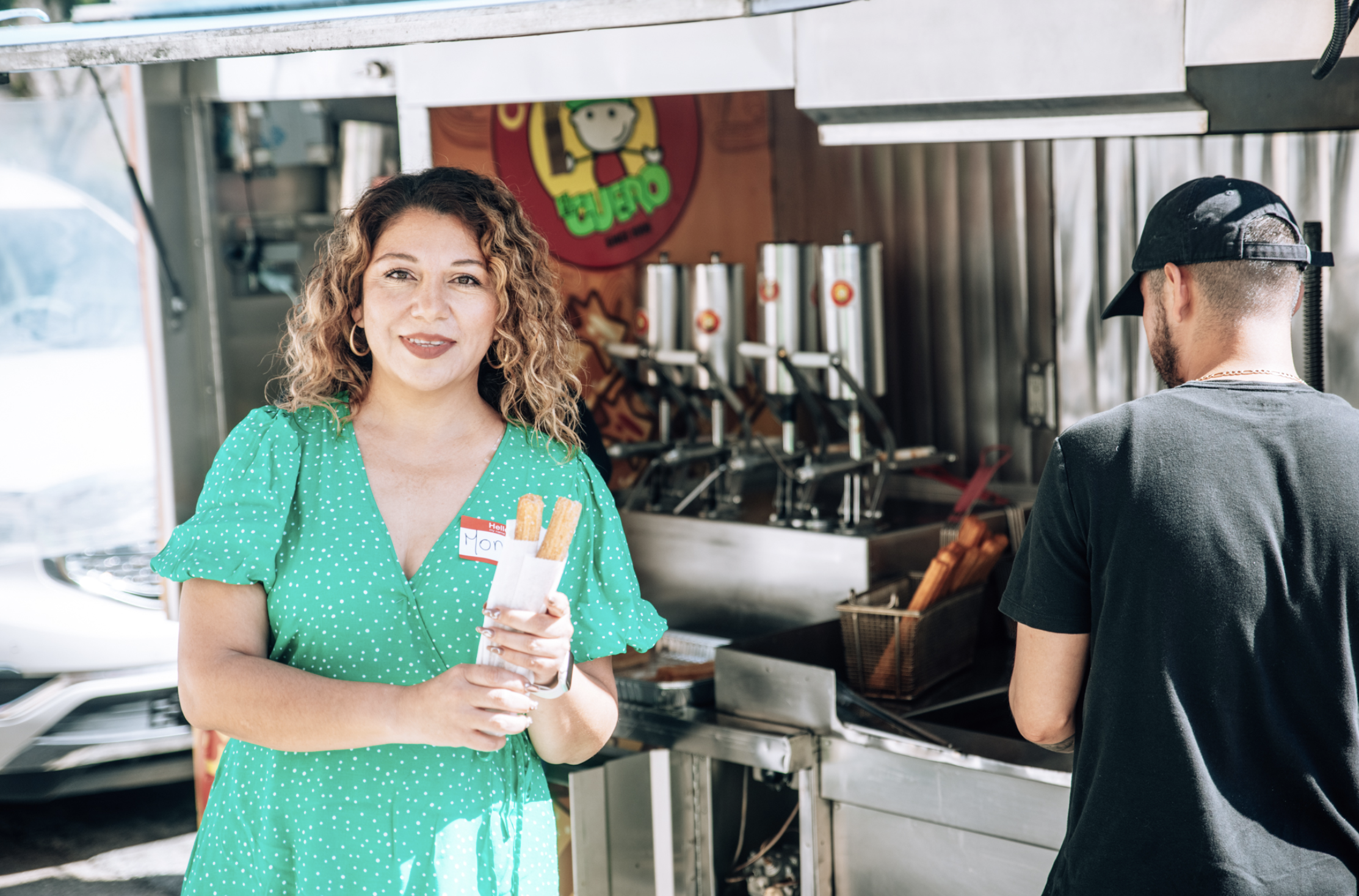 A woman with curly hair wearing a green dress holds two churros at a food stand, with another person in a black t-shirt and cap nearby, in an outdoor setting.